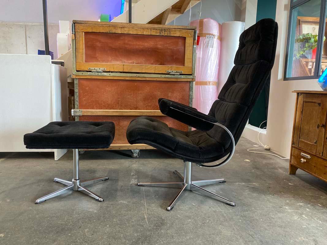 Designed in 1960, this lounge armchair is the perfect seat for those who like to rest their feet from time to time. The armchair with matching stool is upholstered and decorated with buttons. The swivel feet are chrome plated. The black corduroy