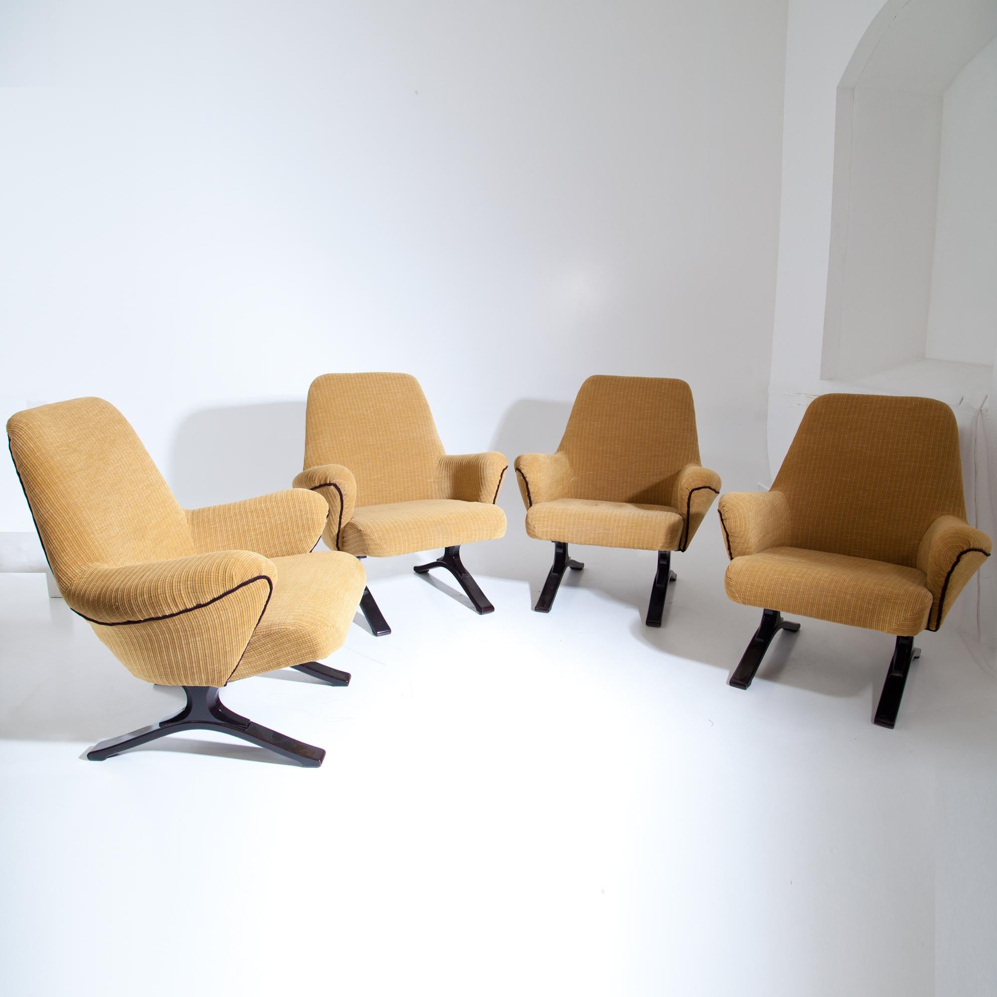 Four lounge chairs on dark stained feet with trapezoidal backrests, deep seats and armrests. The chairs are covered with a yellow textured fabric with black piping.