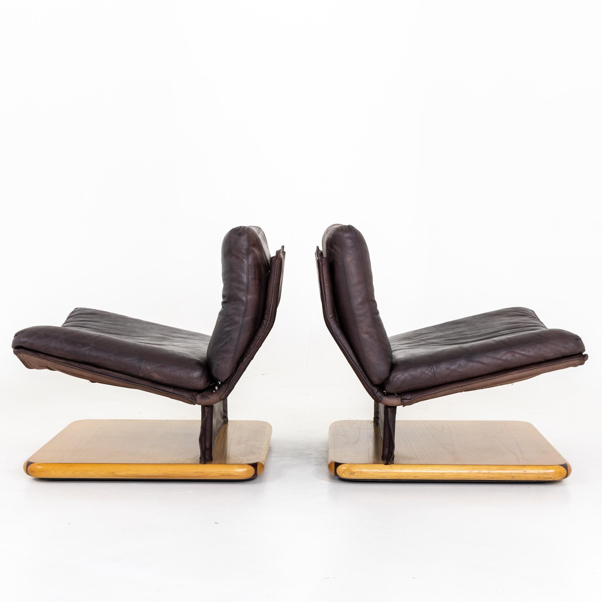 Pair of lounge chairs on rectangular veneered bases and seat covered with brown leather. Inscribed on the bottom: dipo, S. Omero-Teramo-Italia, mod. Gionata. Signs of age and use.