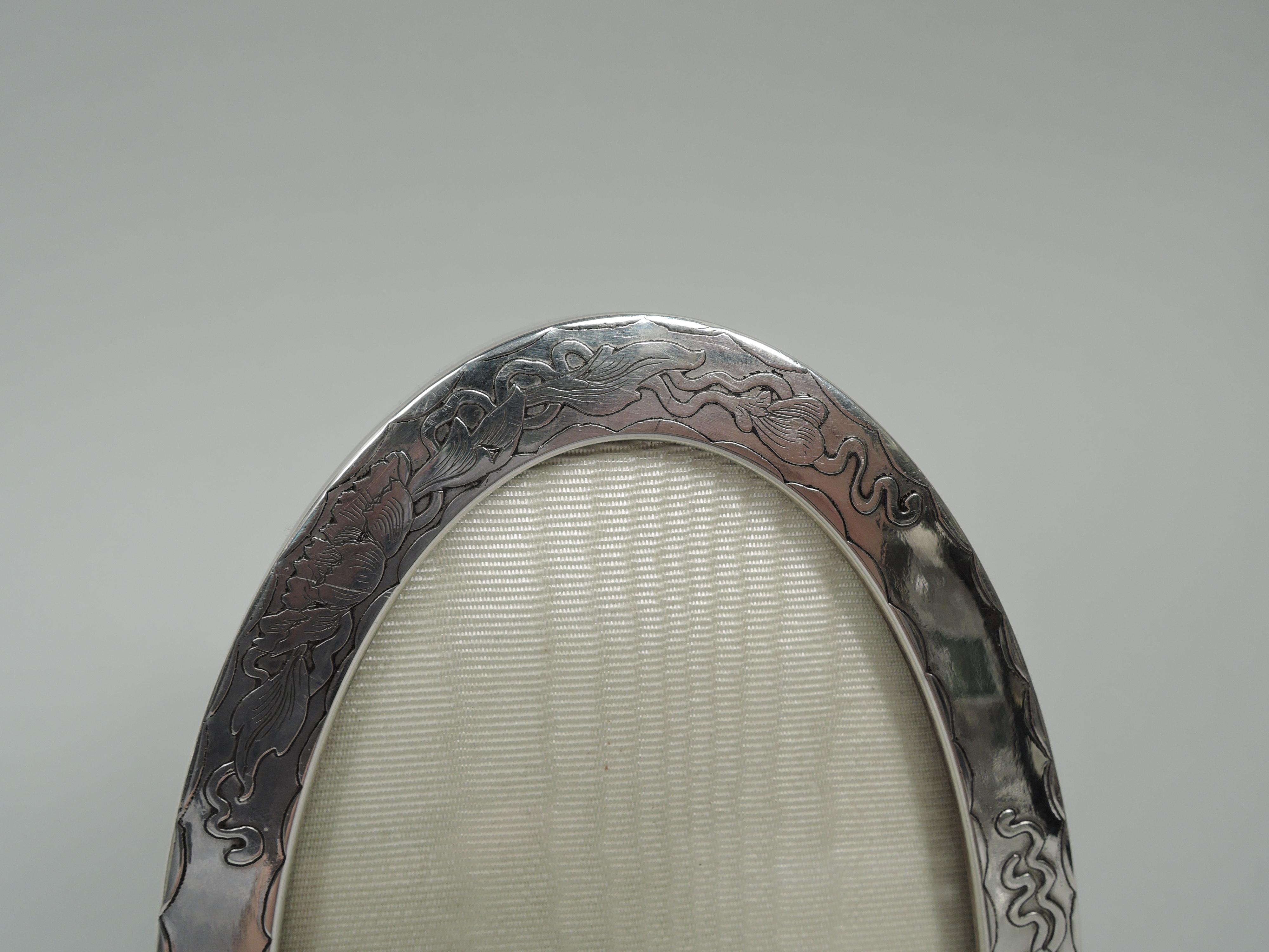 Lovely Art Nouveau sterling silver picture frame. Oval window in same surround. On front acid-etched ornament in form of fluid flower heads and tendrils between scalloped borders; sides plain. Two charming ball supports. With glass, silk lining, and