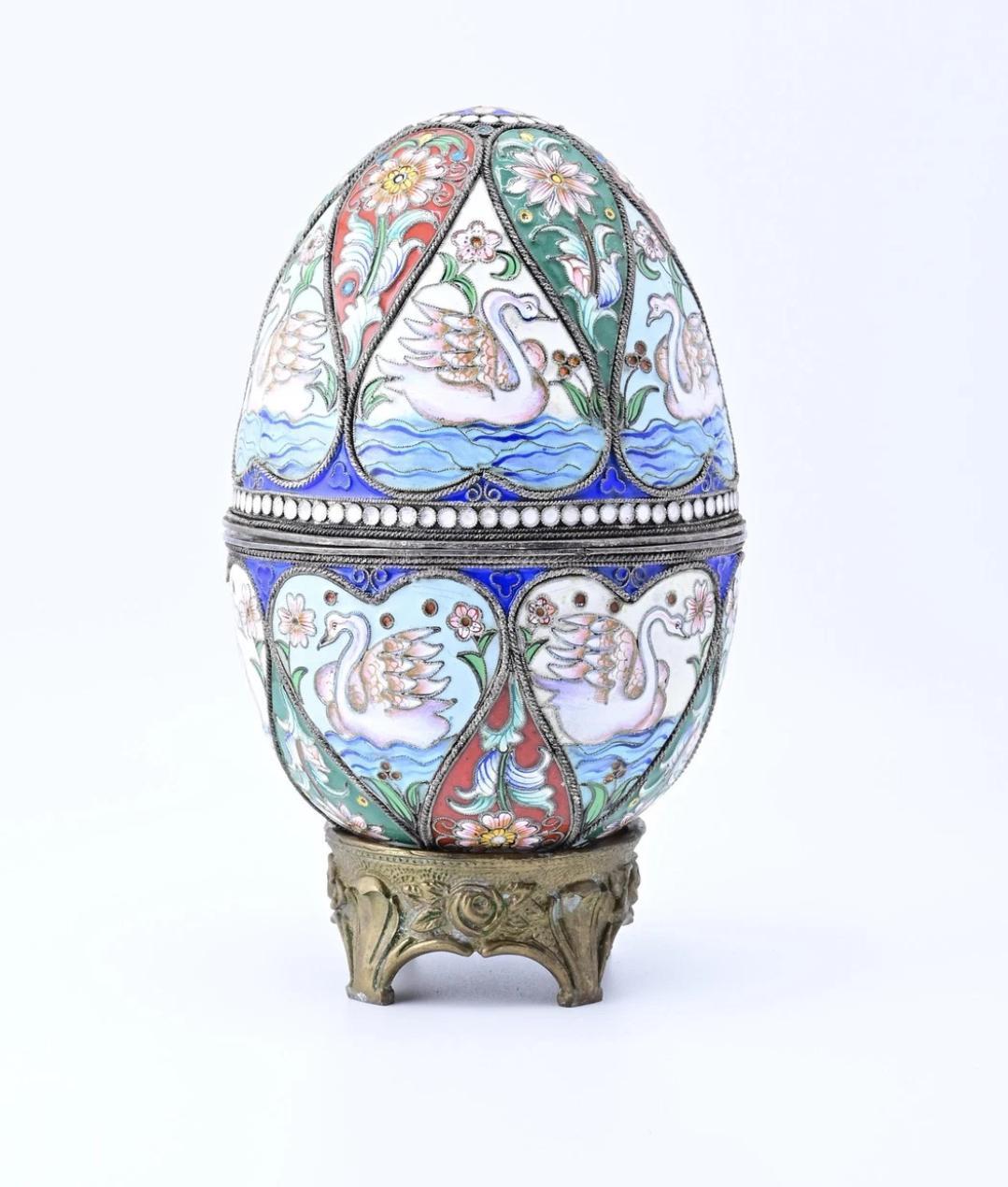 Se trata de un huevo decorativo de plata esmaltada rusa de gran belleza. El huevo presenta una serie de paneles adornados con motivos de cisnes, sobre un fondo de vibrantes azules, verdes y rojos. Cada panel está ribeteado con delicados motivos