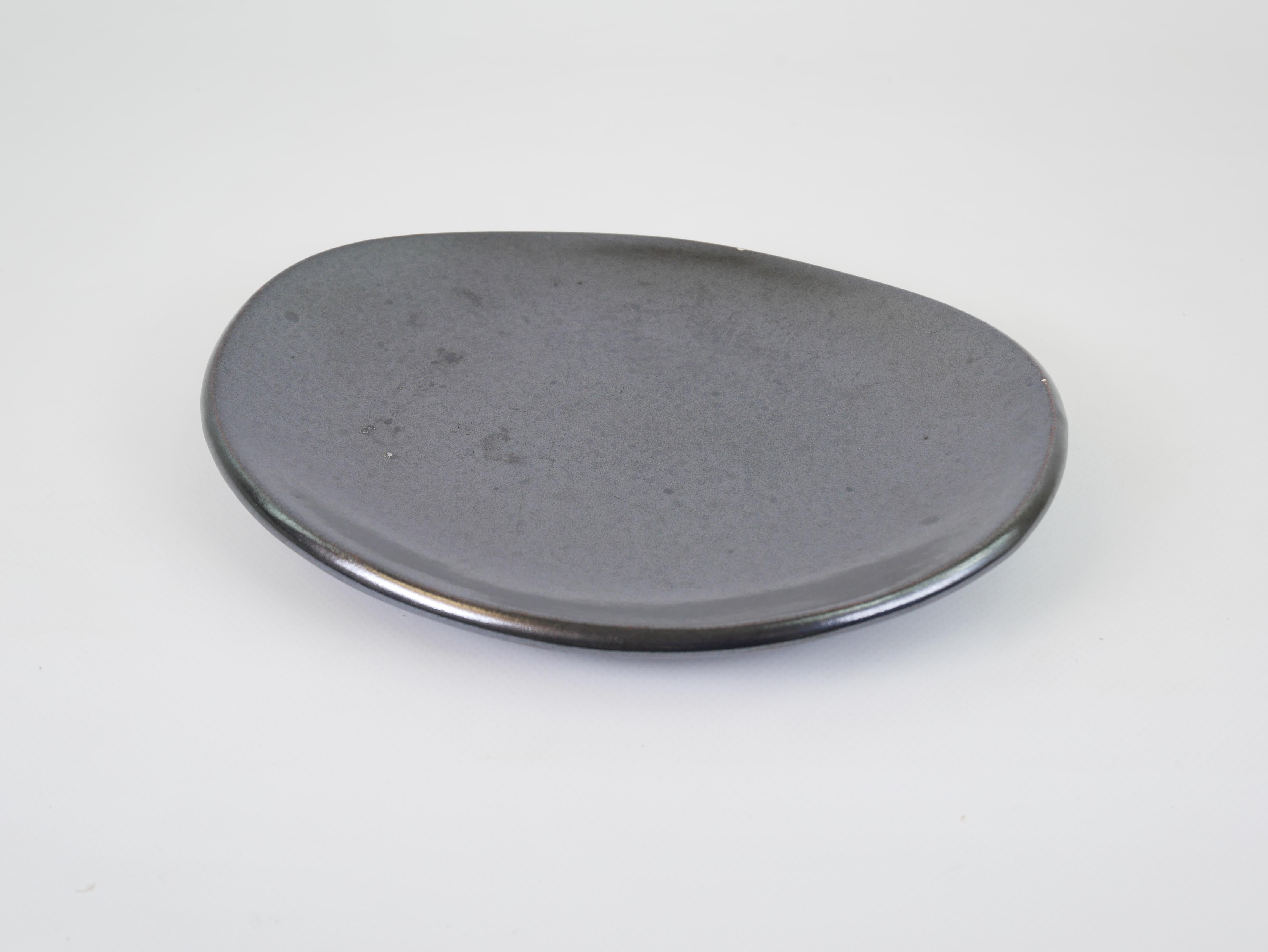 This elegant black glazed ceramic plate showcases the refined craftsmanship of the renowned Madoura workshop. With its soft asymmetrical form and deep, lustrous black glaze, the piece exudes a minimalist yet organic aesthetic. The surface has subtle