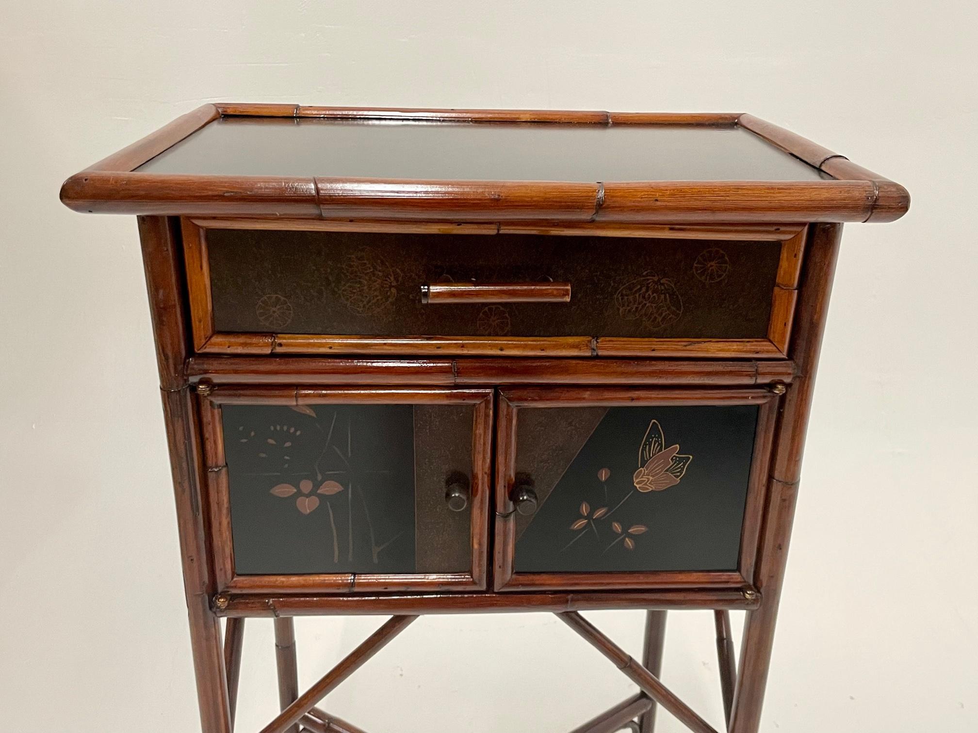 A lovely refined bamboo and chinoiserie decorated ebonized side table or night stand having single drawer over pair of doors that open to storage within. Legs are slightly splayed with Asian flair and x stretcher.