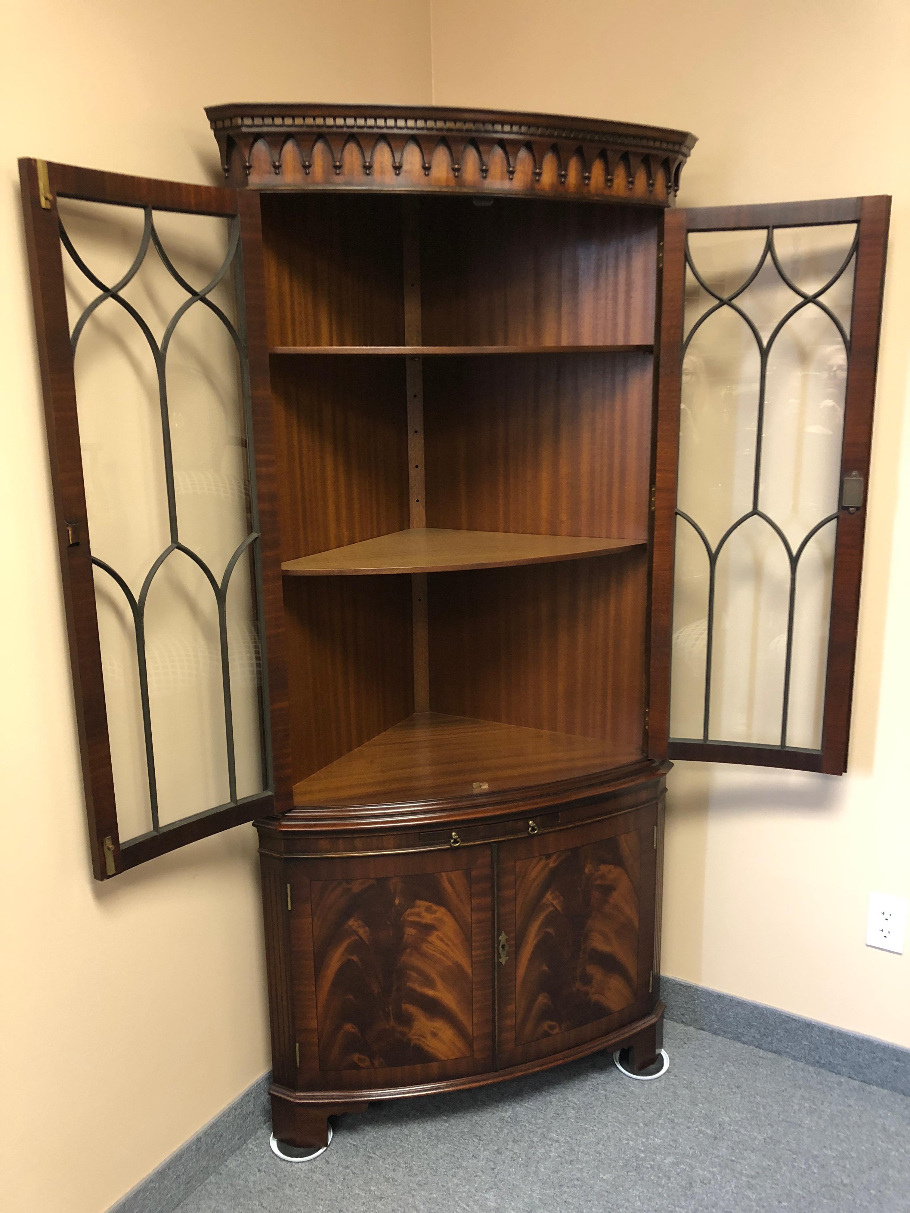 Beautifully made flame mahogany corner cupboard having a curved front with two lovely paned glass doors with 3 interior shelves, gorgeous carved wood decorative pediment, a pull out glide, and two doors at the bottom with storage inside. Key