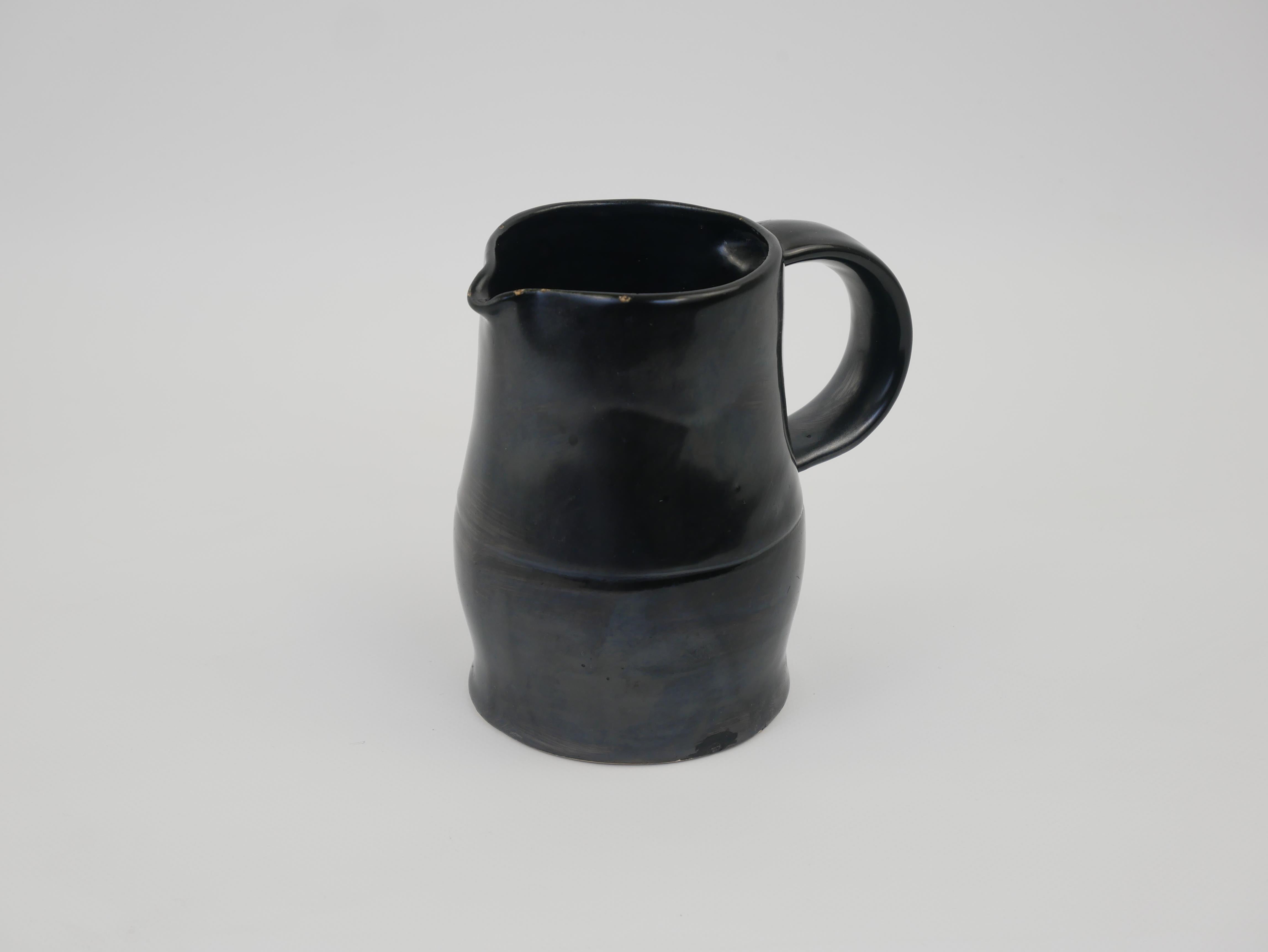 Il s'agit d'un petit pichet en céramique noire datant des années 1950, fabriqué à Vallauris, une ville du sud de la France réputée pour son art de la céramique. La pièce présente une forme simple mais élégante, avec un corps arrondi, un col