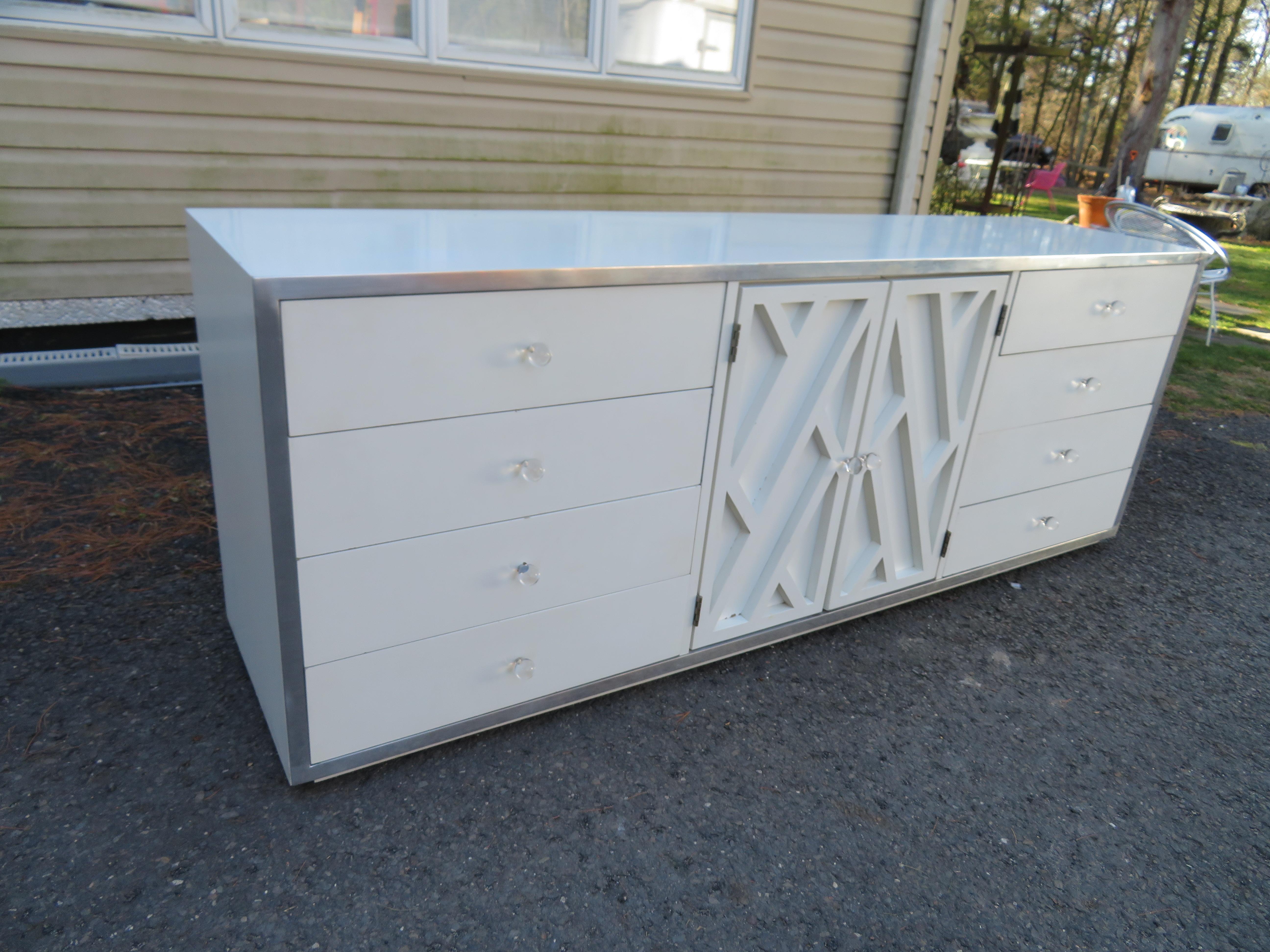 Lovely Milo Baughman-style white lacquered credenza cabinet. Asian-inspired lattice relief detailed doors with lucite pulls open to reveal two deep drawers. A bank of 4 drawers each on either side is all surrounded by a shiny aluminium frame. This
