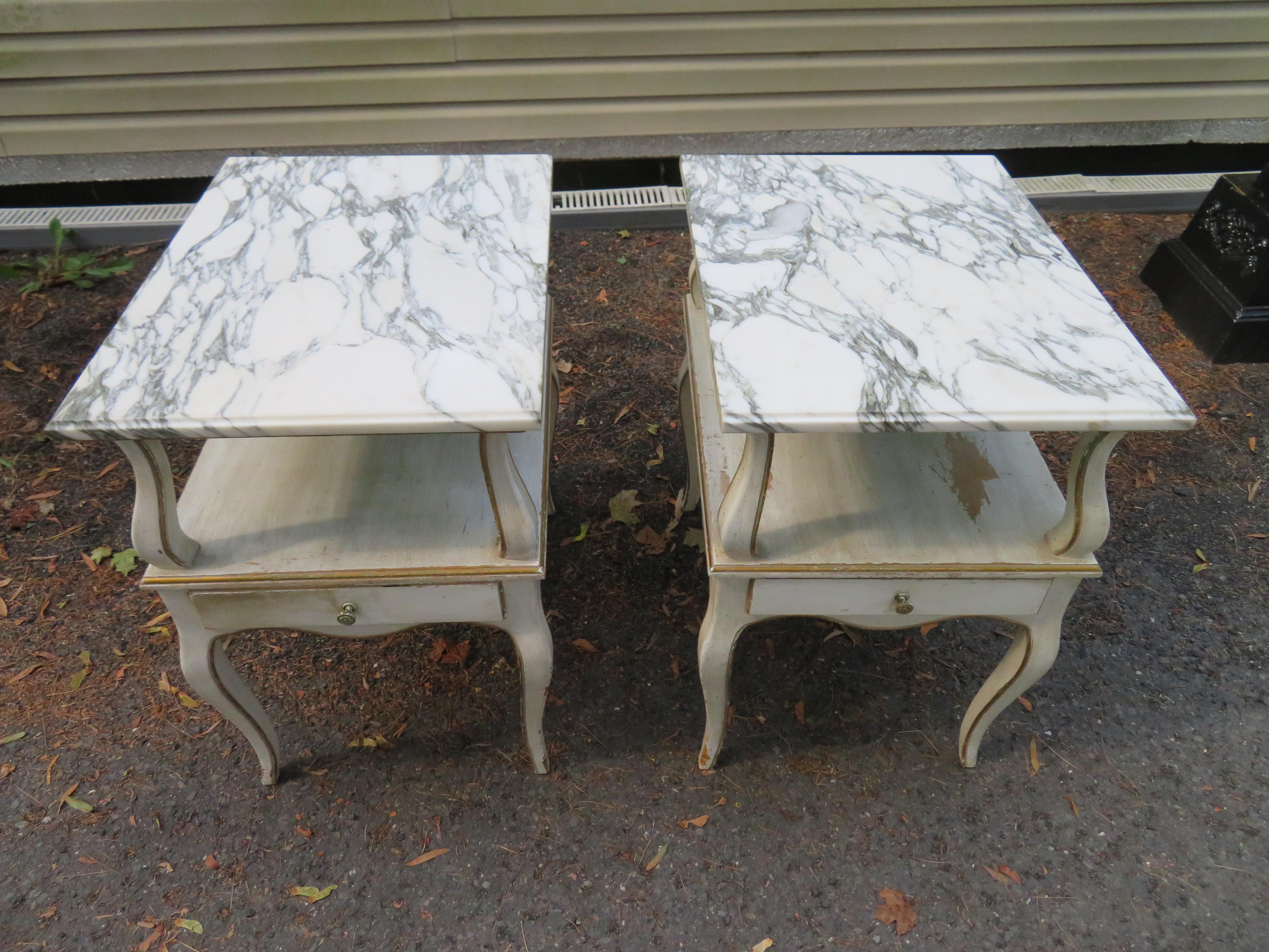 Lovely pair of Dorothy Draper style marble-top nightstands with one-drawer each. The original creamy white finish is intentionally distressed and has gold accents.