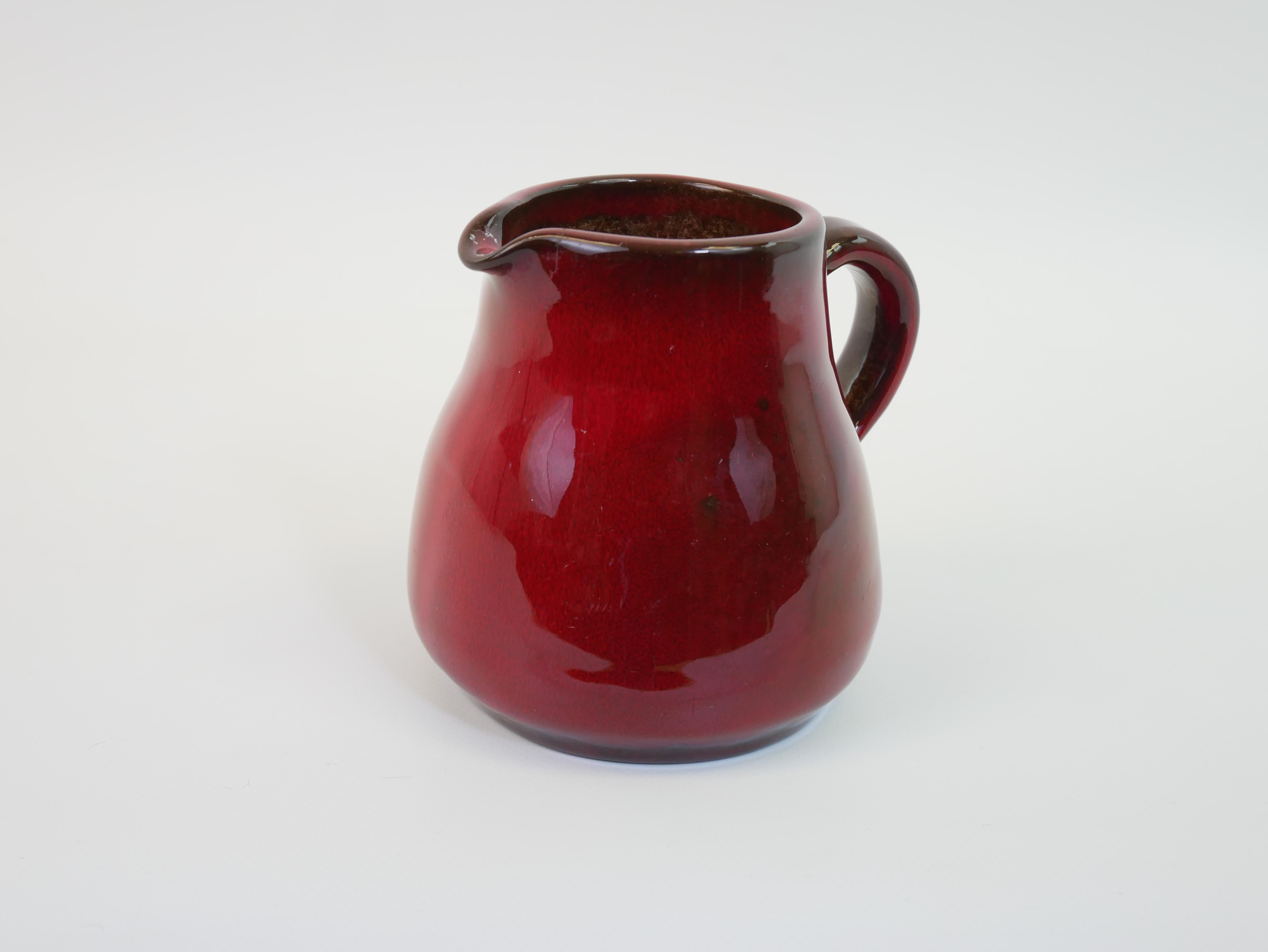 Ce charmant pichet de 12 cm de haut, qui rappelle le style emblématique de Georges Jouve en matière de céramique, présente un émail rouge cerise exquis qui séduit par sa profondeur et sa richesse. La glaçure brillante reflète magnifiquement la