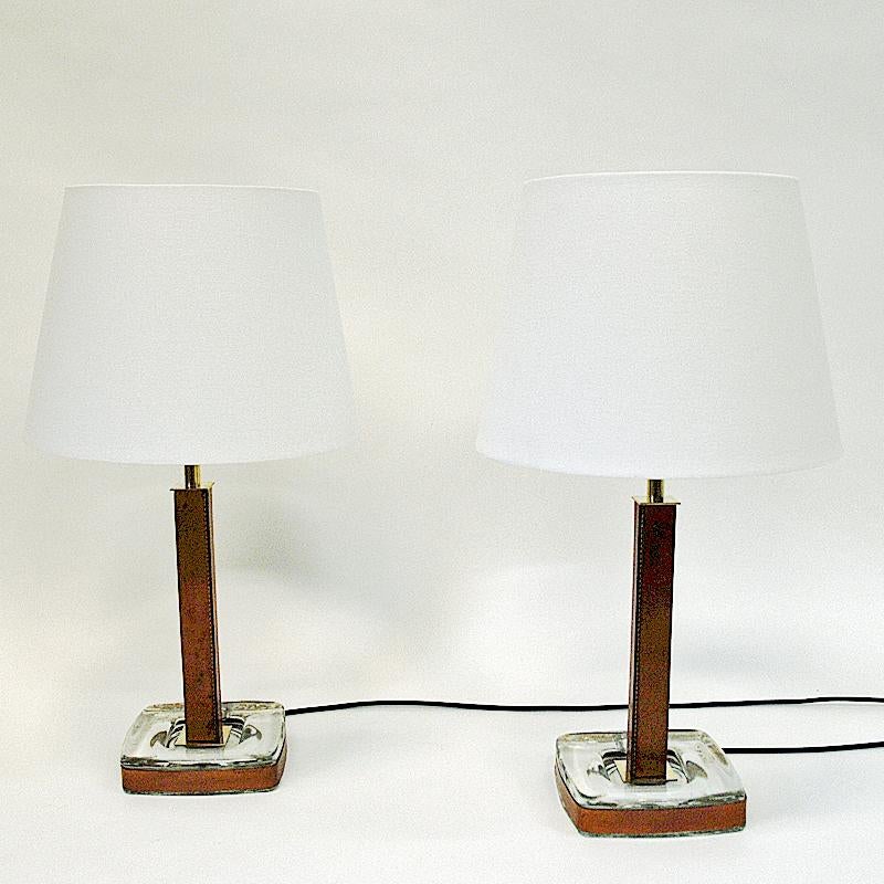 Nordic and special shaped pair of table lamps in cognac brown leather stom by Uppsala Armaturer, Sweden 1950s. Beautiful together or as single lamp, on a table or in the window. Clear glass base with leather edges around and black new cords. Brass