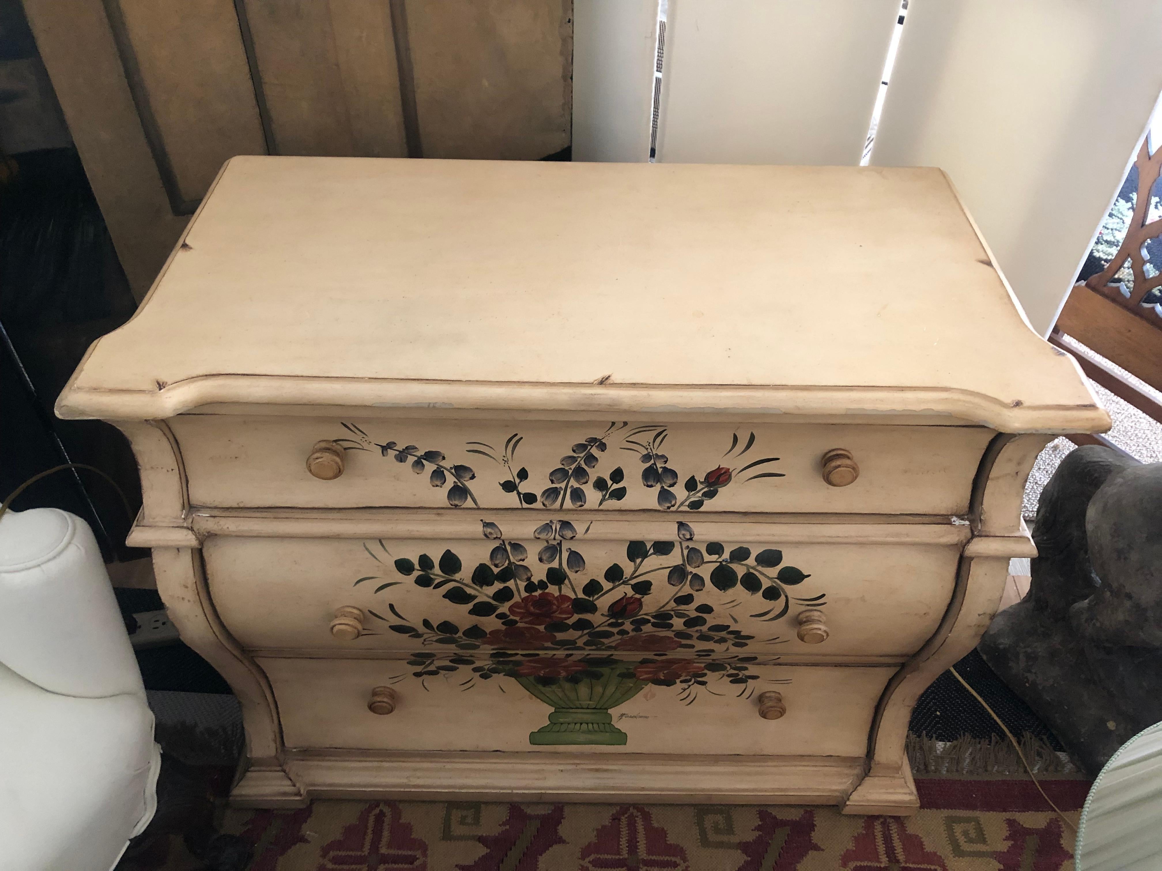 A pretty hand painted vintage medium sized dresser having 3 drawers and a bowed shape. The background is an antiqued white with a floral arrangement in a green urn painted on the front.