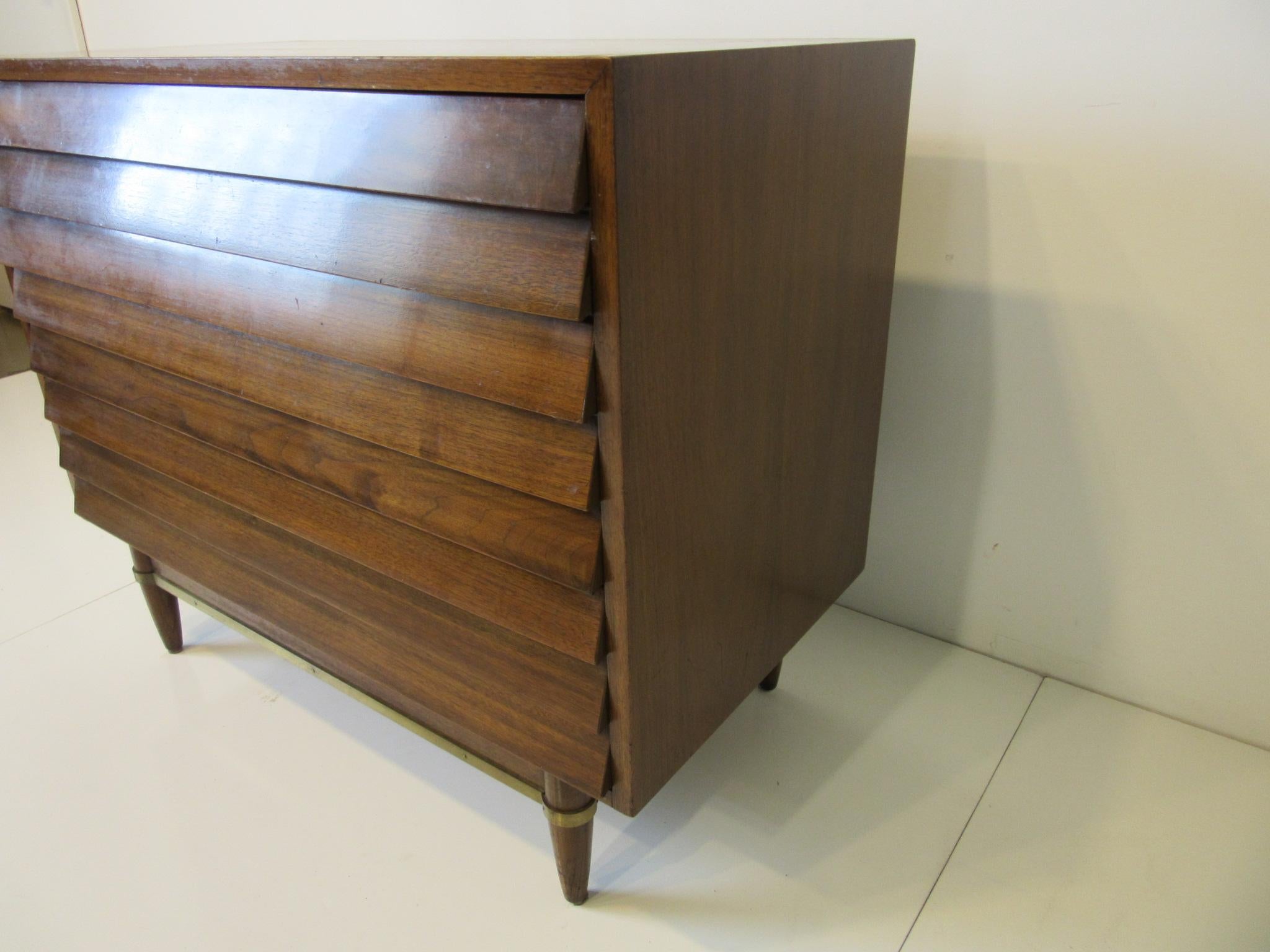 A smaller scale and well constructed walnut dresser/ chest with three large drawers the top one having dividers. The louvered front drawers gives this piece a high style sitting on conical legs with brass stretchers manufactured by American of