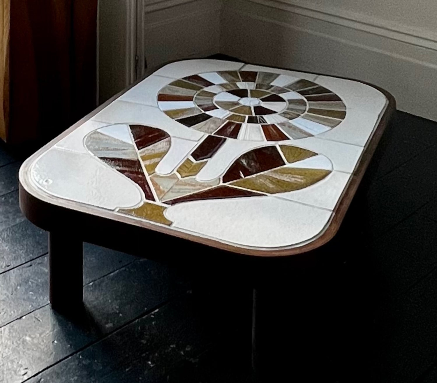 A ceramic coffee table by the French ceramicist Roger Capron. Glazed tiles in a palette of greens and browns form the modernist outline of a flower, set against a lighter neutral background. The heavy table legs are wood and the top is finished with