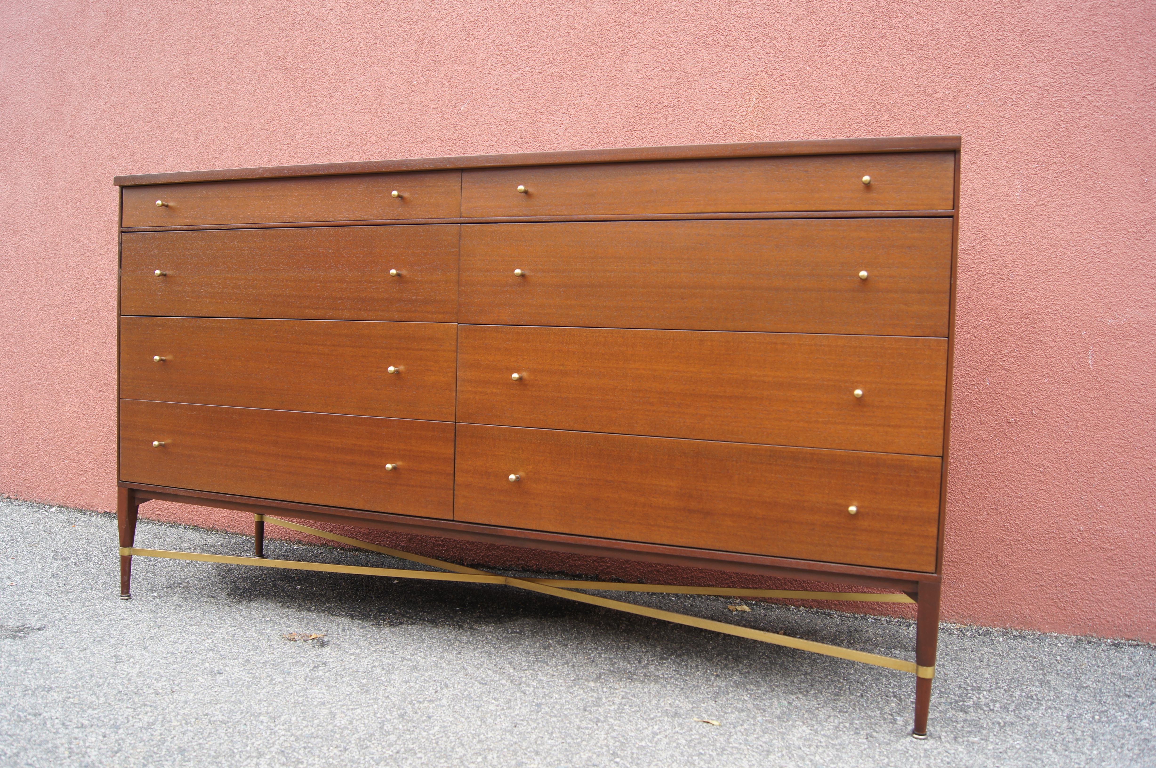 Paul McCobb designed this double dresser for the Calvin Group's Irwin Collection. A walnut-stained mahogany case sits on delicately tapered legs with a brass cross brace. The eight drawers, the topmost of which are shallower, feature small rounded