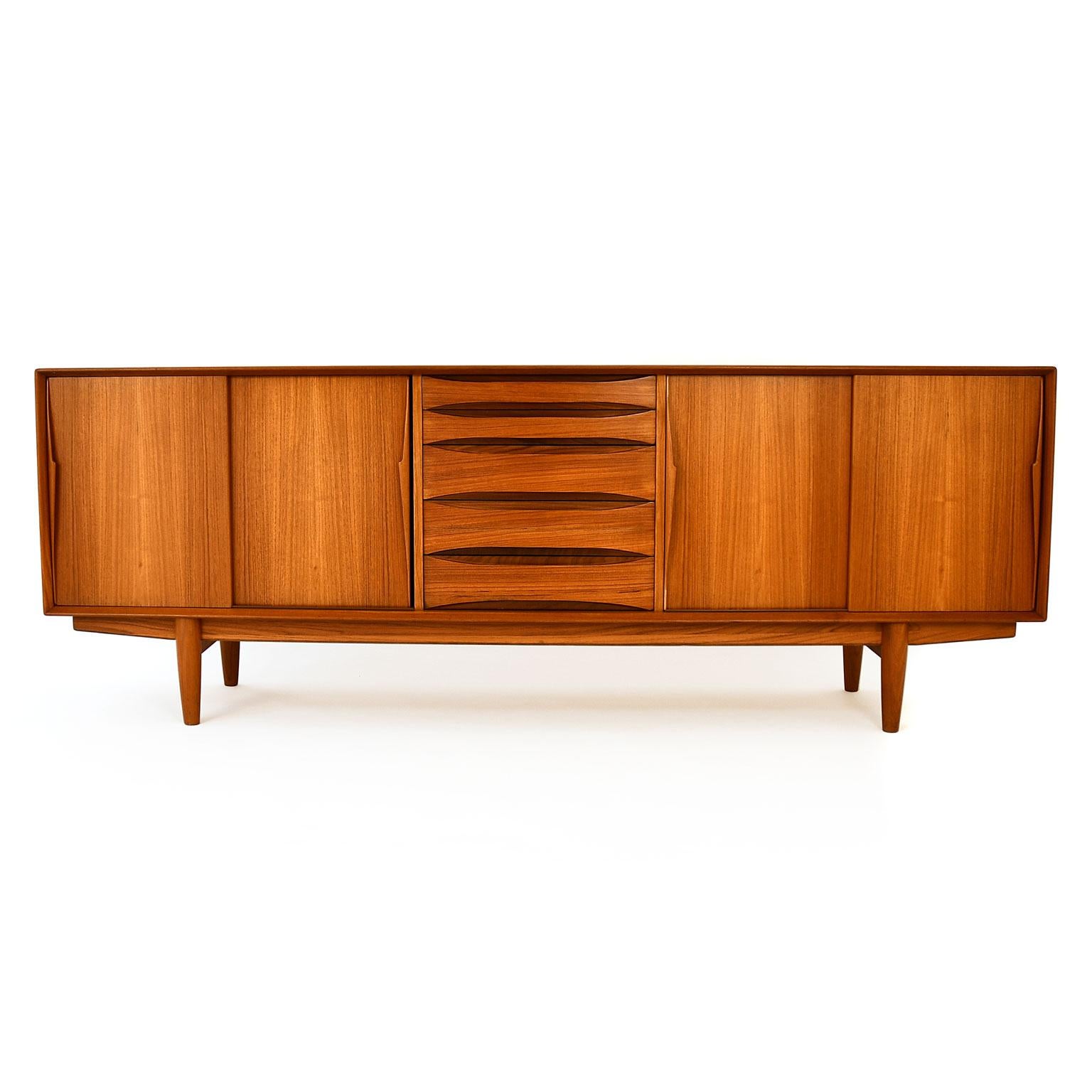 Danish low midcentury sideboard designed by Arne Vodder in the late 1950s. Dyrlund produced it in the 1960s. The frame is made of solid teak wood, and teak wood veneer. Two sliding doors on each side hiding shelves. 5 drawers, 2 smaller and 3 larger