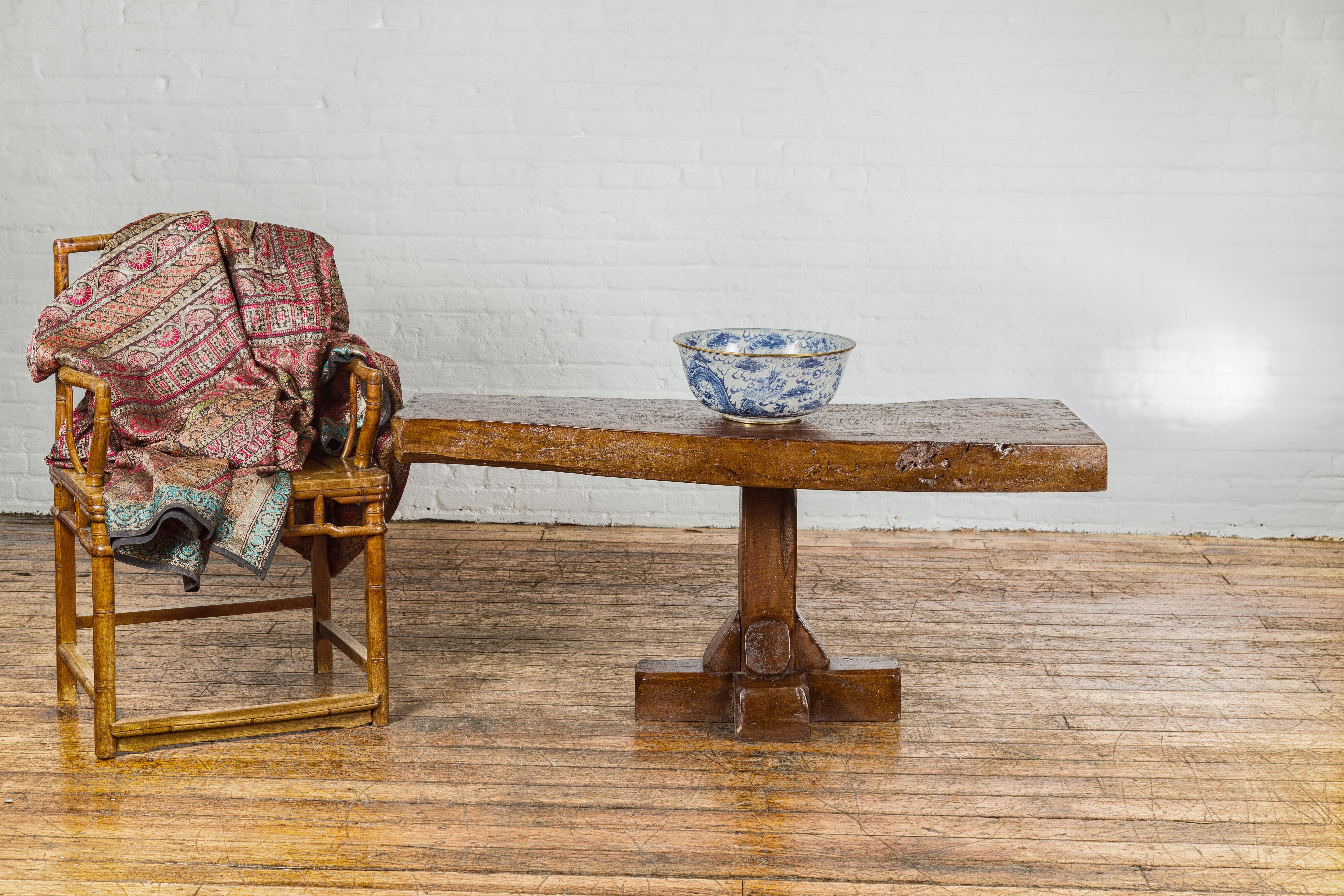 A low wooden console table from the early 20th century with rustic top and pedestal quadripod base. This early 20th-century low wooden console table exudes rustic charm and understated elegance. With its robust and weathered top, the table showcases