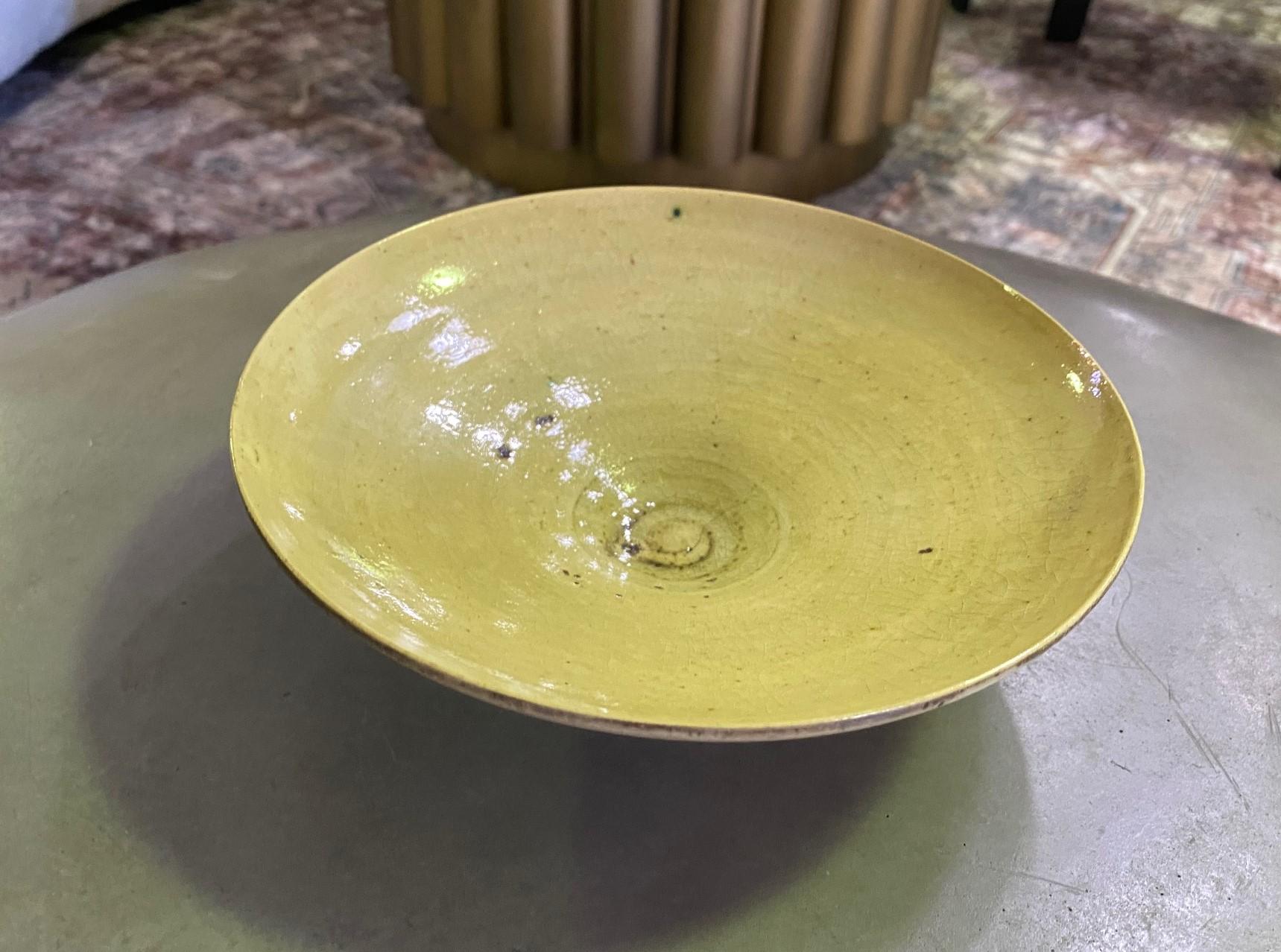 Precioso cuenco con pie de cerámica esmaltada en tonos tierra, amarillo, verde y marrón, obra de la famosa ceramista británica de origen austriaco Lucie Rie.

El cuenco está firmado con el sello impreso de Lucie Rie en la base.

Una obra rara y
