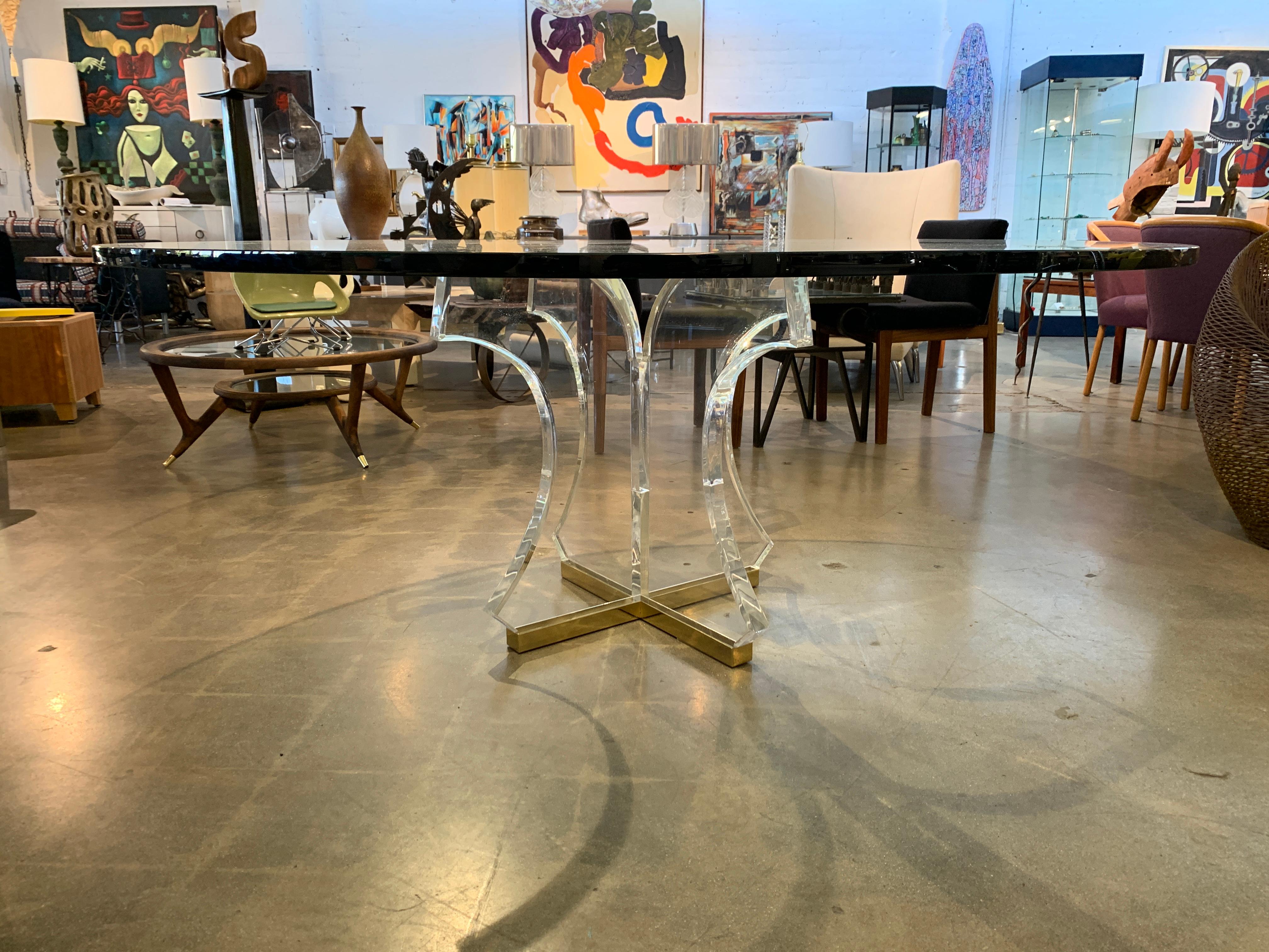 A pretty dining table in the manner of Pace, Bigelow, Spectrum or Charle Hollis jones. It features a brass or plated base with curved heavy and thick Lucite support columns. The glass is 1 inch thick and 60 inches in diameter. The glass has a few
