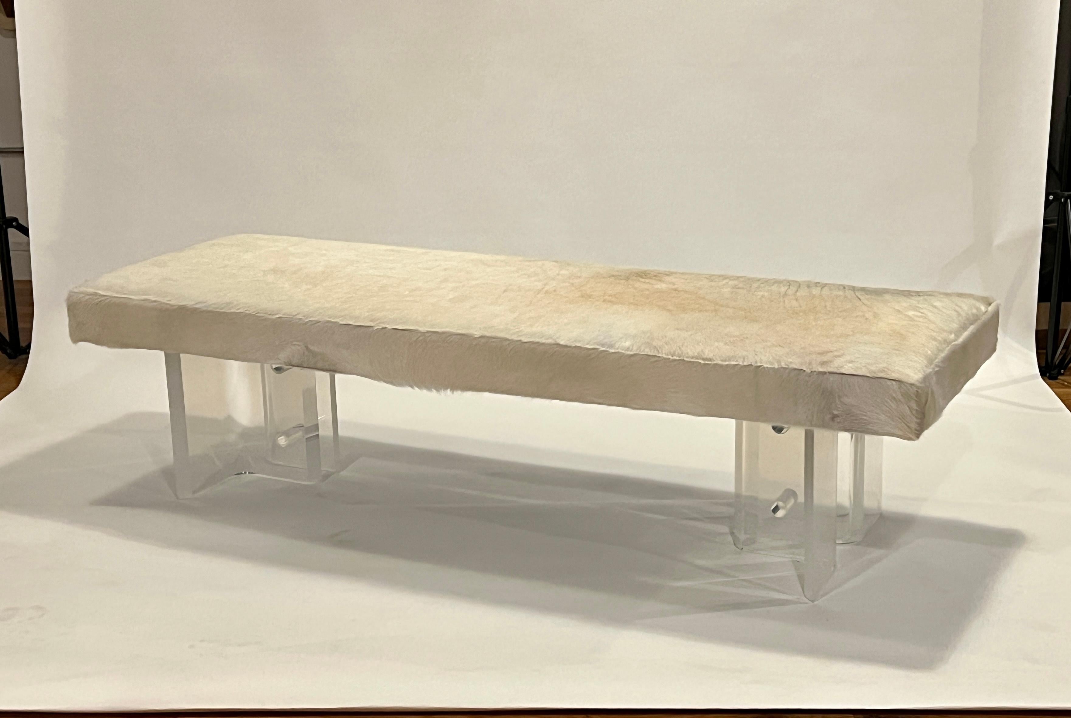 Midcentury lucite table bases have been reimagined into a long bench freshly upholstered in blonde cowhide. 
This sturdy and well-crafted bench features a box stitch seat and decorative lucite bases. A thick platform was installed to prevent bowing