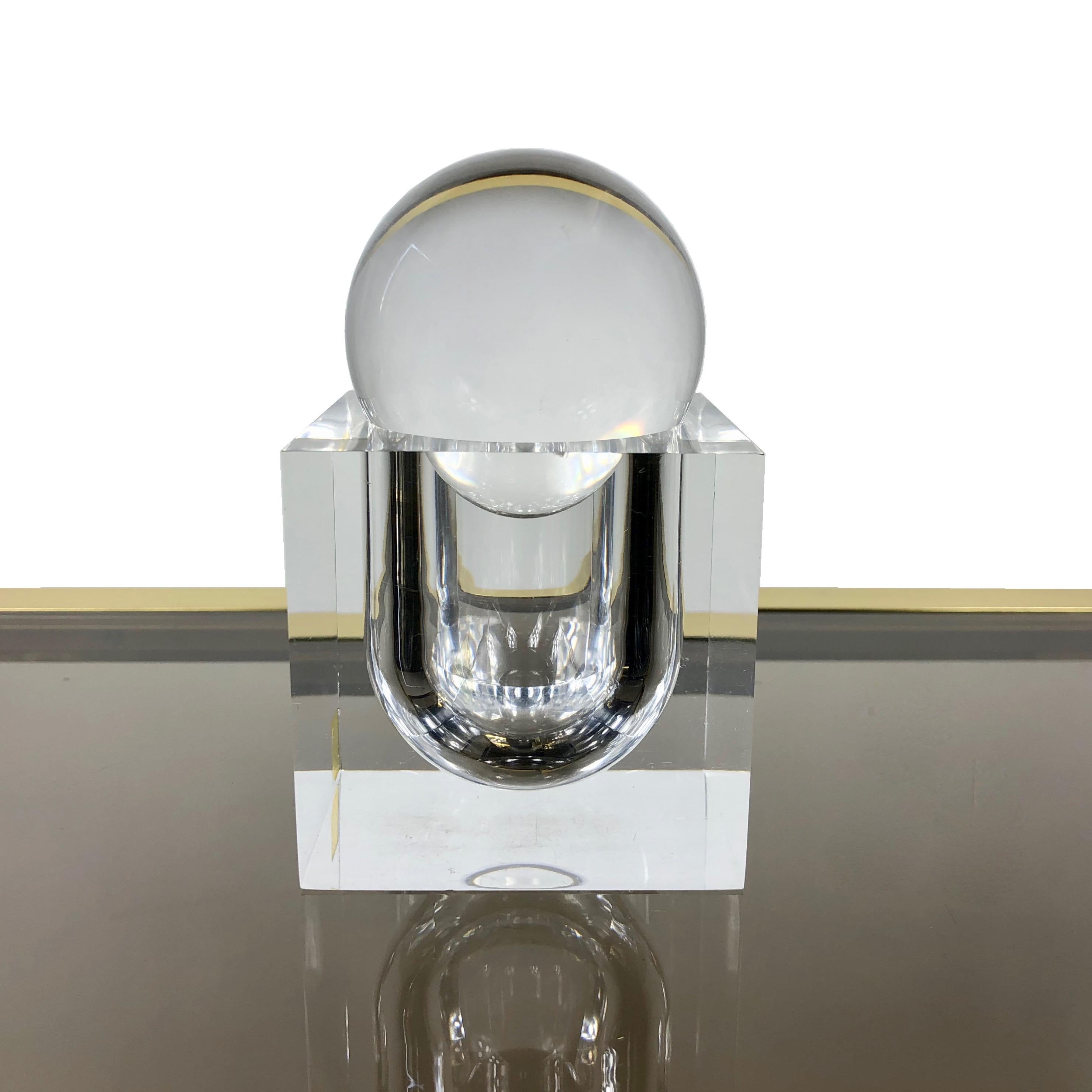 This Lucite art sculpture by Team Guzzini consists in two pieces: a sphere and a square base featuring a slot in which the sphere is inserted and which can also be used as a container. It can also be used as a paperweight or as a little box. 
The