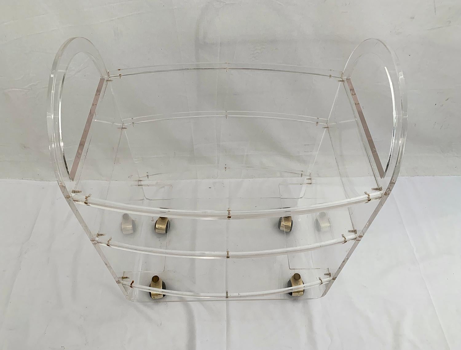 This Lucite trolley rests on four wheels finished by a brass hull. It has three shelves, one shorter for the bottle space. A small piece of the ruler on the side of one of the tablets is missing.

Ce chariot en plexiglas repose sur quatre