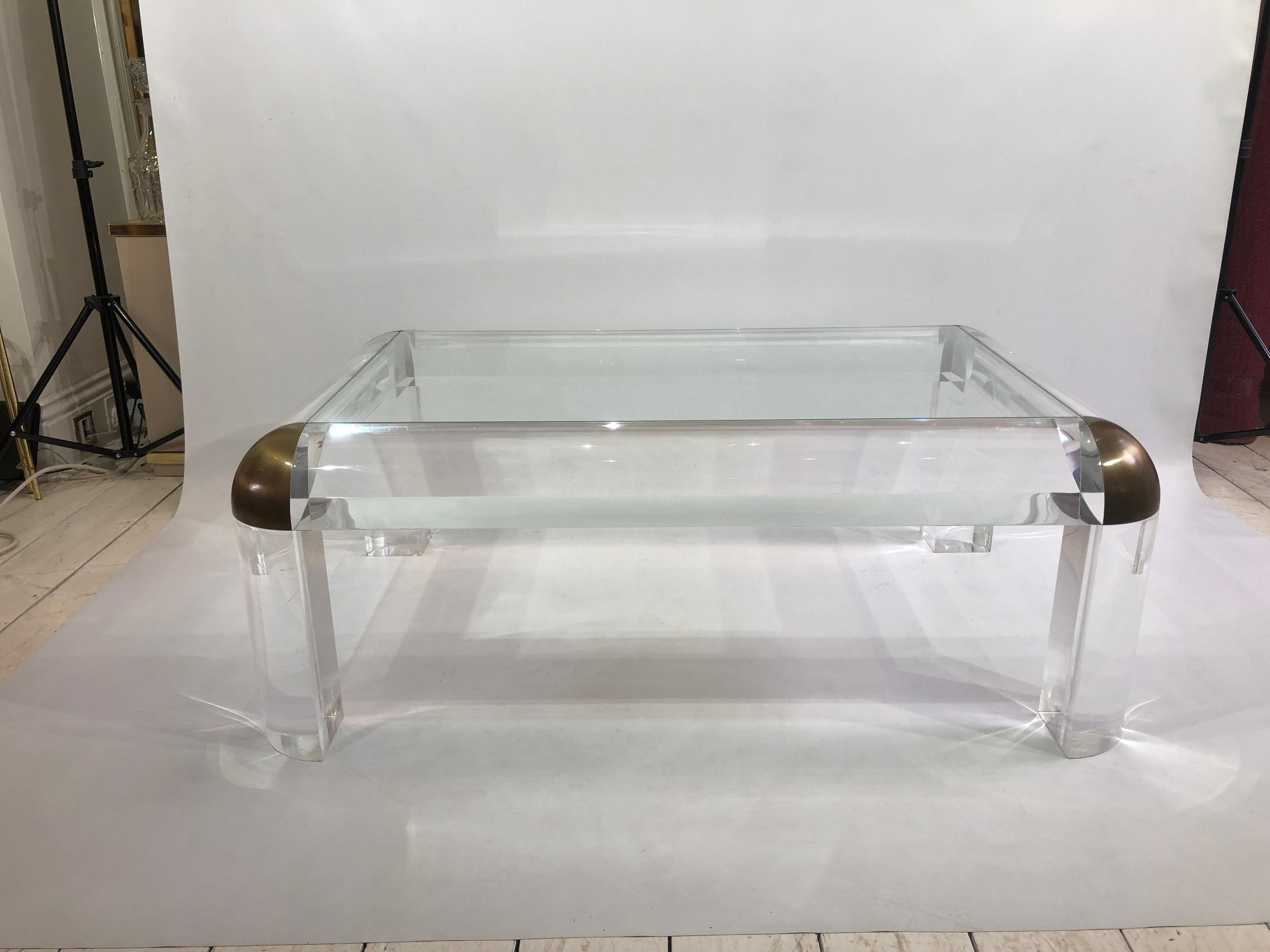 This amazing lucite and brass coffee table, very much in the style of Karl Springer, was imported from the United States. Four sturdy legs house a clear glass top, and at each edge you find a curved brass join in an antique finish. 

This is an