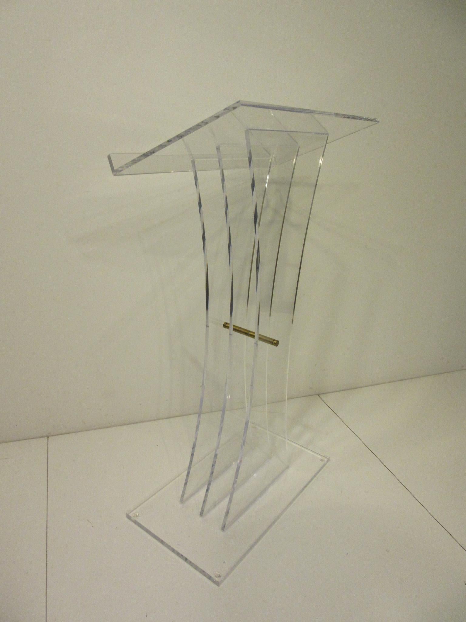 A very sculptural Lucite and brass lectern podium or hostess stand, this high end piece has three graduating uprights with a work surface having a stop to the bottom edge of the surface. The stand has a polish brass rod holding the uprights together