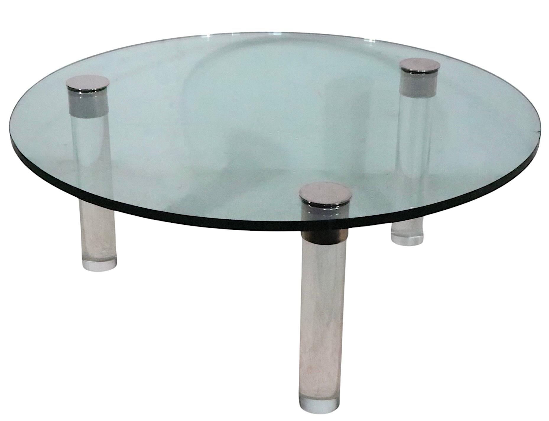 Table basse de style Hollywood Regency avec un plateau en verre trempé épais, monté sur trois pieds en lucite solide, avec des garnitures chromées. La table est en bon état d'origine, présentant un petit défaut dans le plateau en verre -.  ébréchure