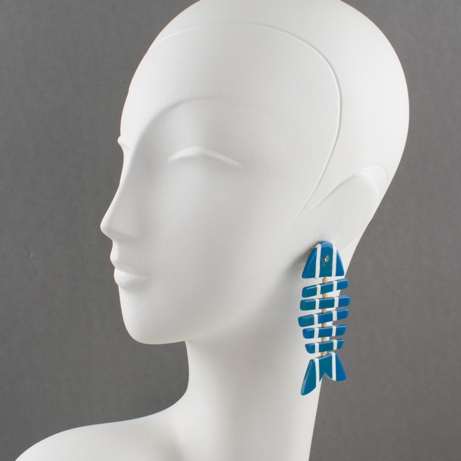 These stunning Lucite clip-on earrings feature a dangling geometric fishbone shape in a blue and white striped pattern ornate with crystal rhinestone eye and white glass bead spacers. There is no visible maker's mark.
Measurements: 3.19 in long (8