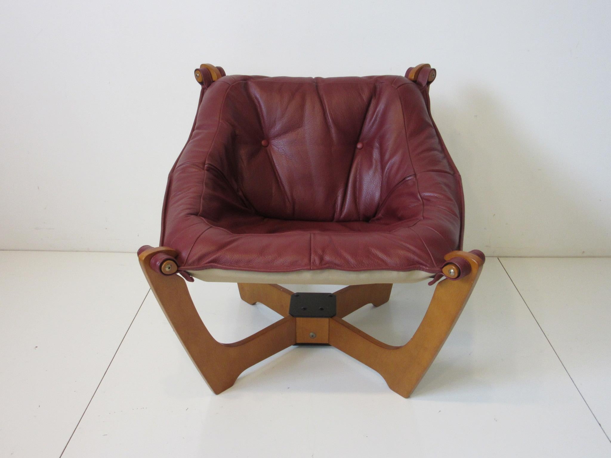 A lounge chair in a deep rich burgundy colored leather with X crossed wooden base which holds each corner of the seat in a sling style arrangement. The corners are securely held together with brass bolts and wood, well-crafted and super comfortable