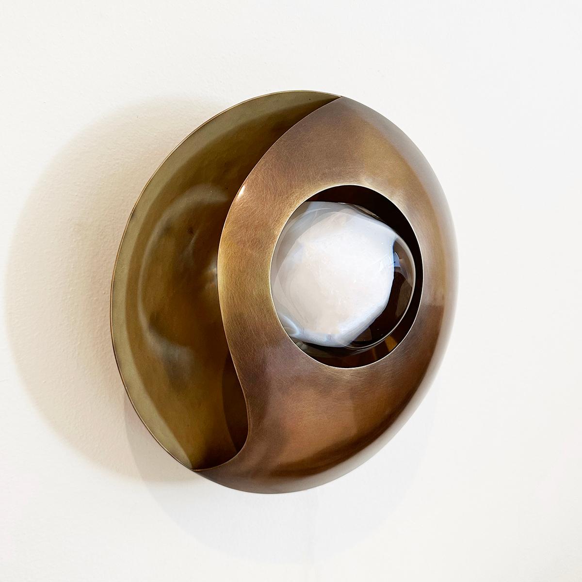 The Luna wall light’s simple round shape is elevated by its elliptical cross-section and distinctive cut-outs that let out a soft glow. At the center is our Sfera glass handblown in Murano. Shown with a satin brass interior and bronzo nuvolato