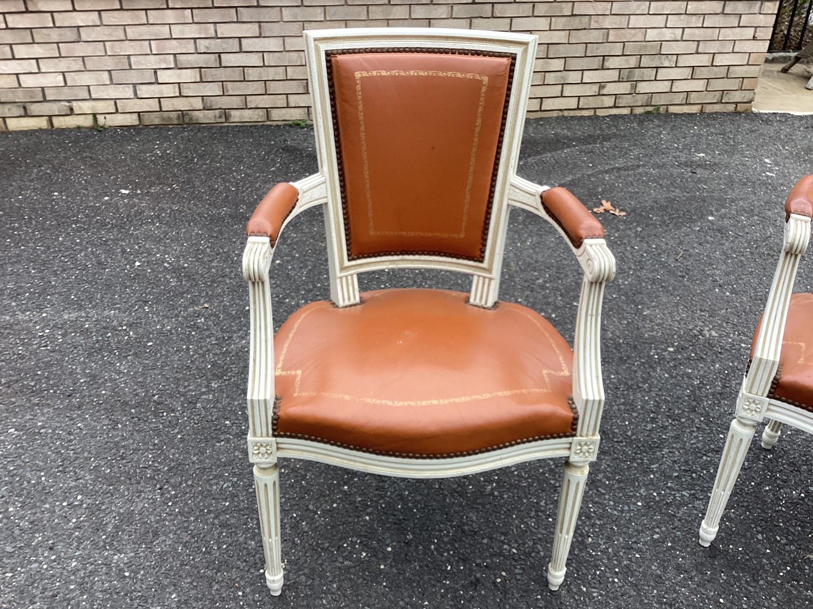 Classic pair of painted white Louis XVI style leather arm chairs featuring a square back with scalloped seat, straight fluted legs and carved floral motifs at the top of the legs. The upholstery is genuine cowhide leather in a caramel tone with gold