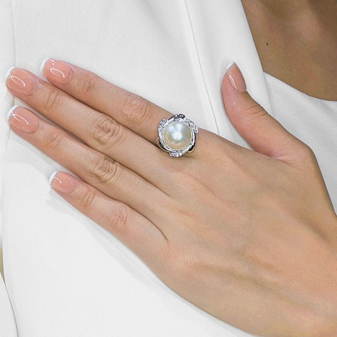 Cette bague d'apparat exsude la sophistication avec en son centre une perle lumineuse de 13,4 mm de couleur Brilliante, encadrée par 9 diamants taille brillant (0,18ct, couleur F-G, pureté VS) qui étincellent à chaque mouvement. Réalisé en or blanc
