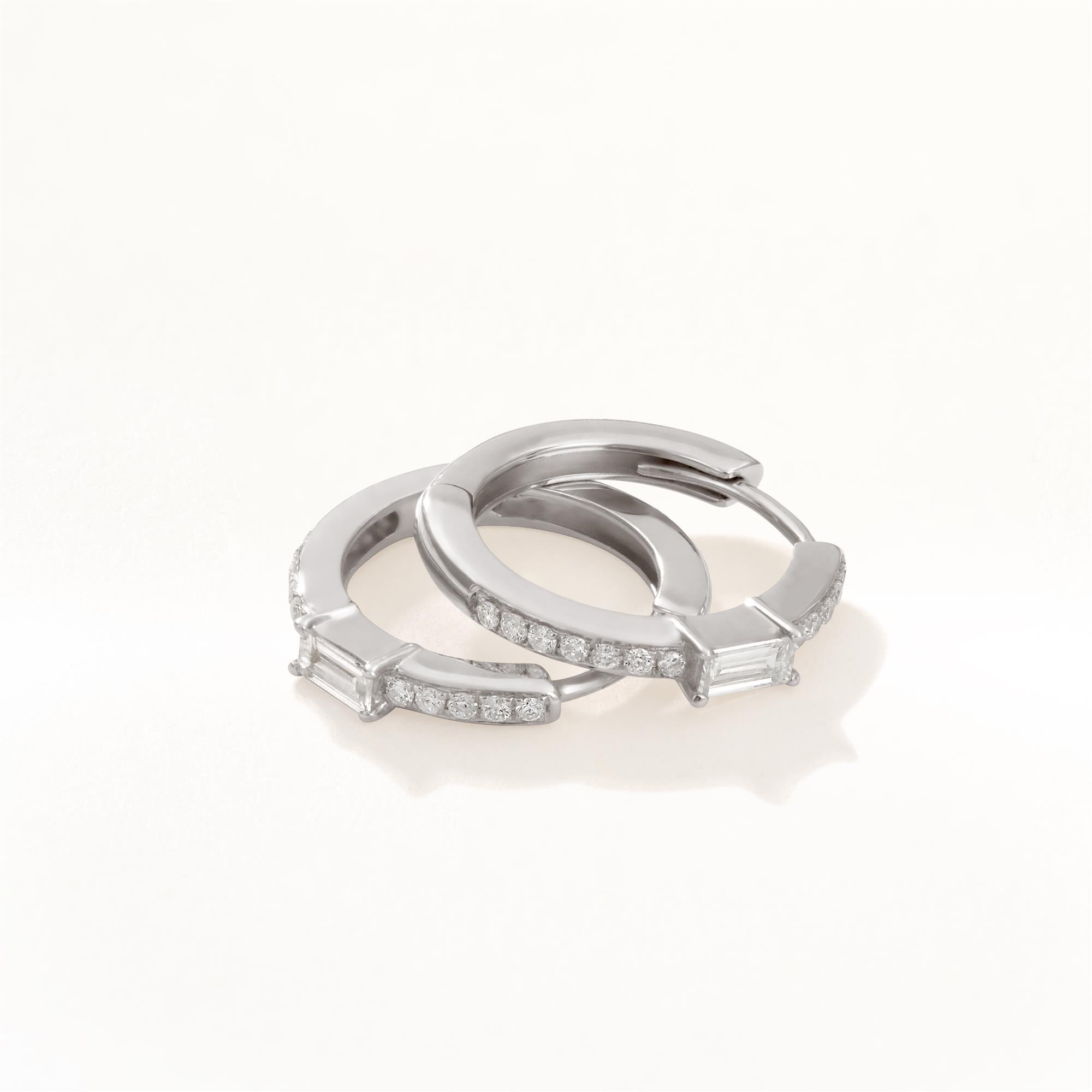 These eye catching luxle diamond huggie earrings crafted in 18k white gold, has a sleek circular design that features a striking baguette-cut diamond at the center, set in prongs for maximum brilliance, above and below the baguette, shimmering