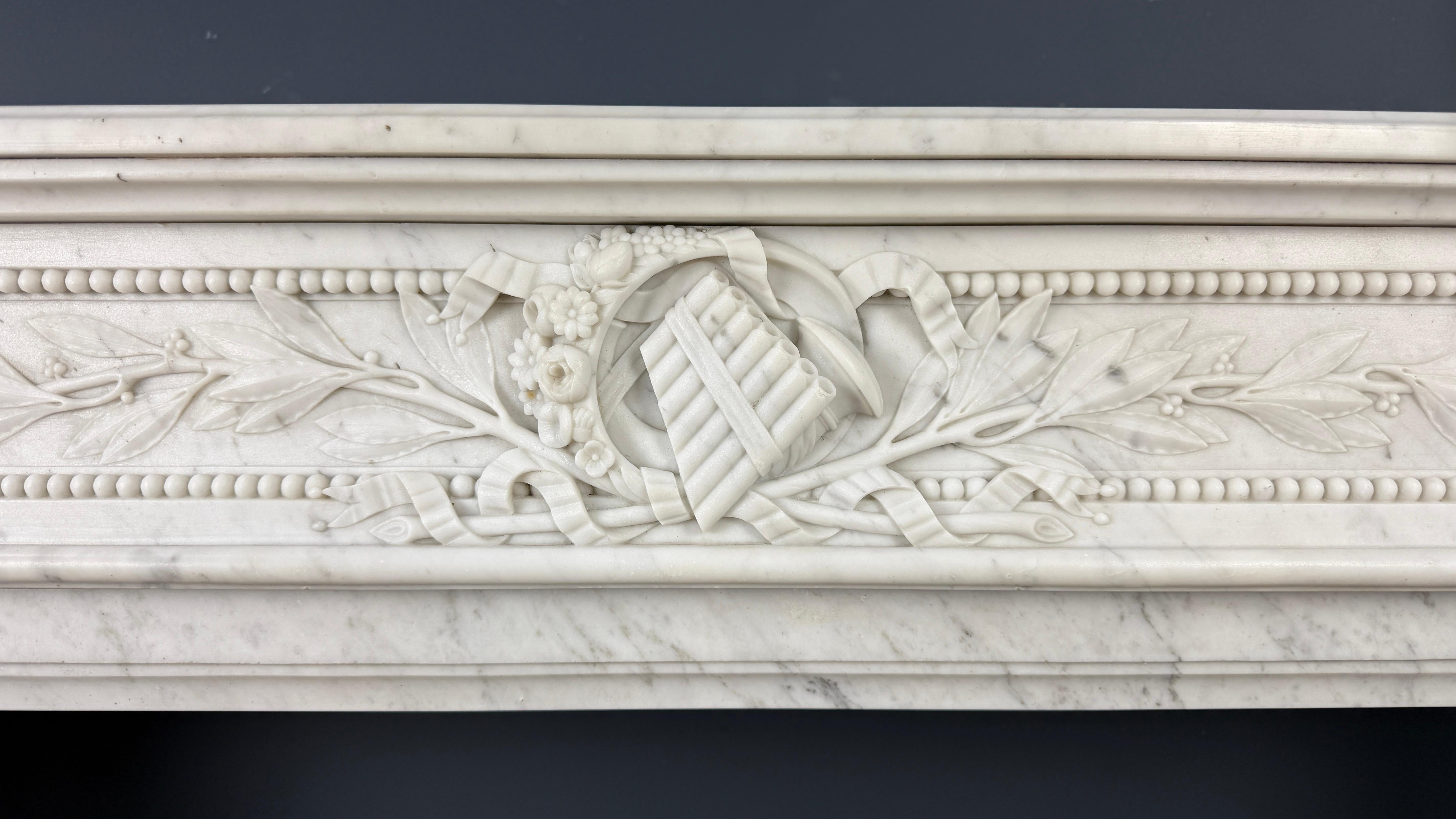 This monumental and richly decorated French antique fireplace mantel is crafted from finely veined white Carrara marble and exudes the classical grandeur of the Louis XVI style. The front is exceptionally detailed, featuring a recessed central