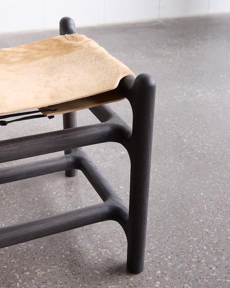 The Lyndoe Bench is a low, sculptural seating piece built around repetition and rhythm. Twin sling seats are suspended within a rounded, solid wood frame that features softly exaggerated joinery and continuous horizontal rails. The dual-seat design