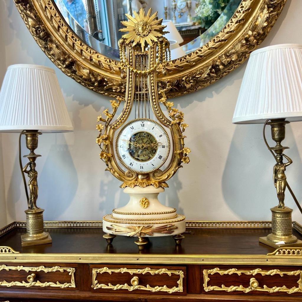 A stunning lyre-shaped clock mounted on a white Carrara marble base, adorned with floral decorations, beads and a sun motif comprising a satyr face in gilded bronze. Its intricate transparent movement called skeleton includes an extra arm for