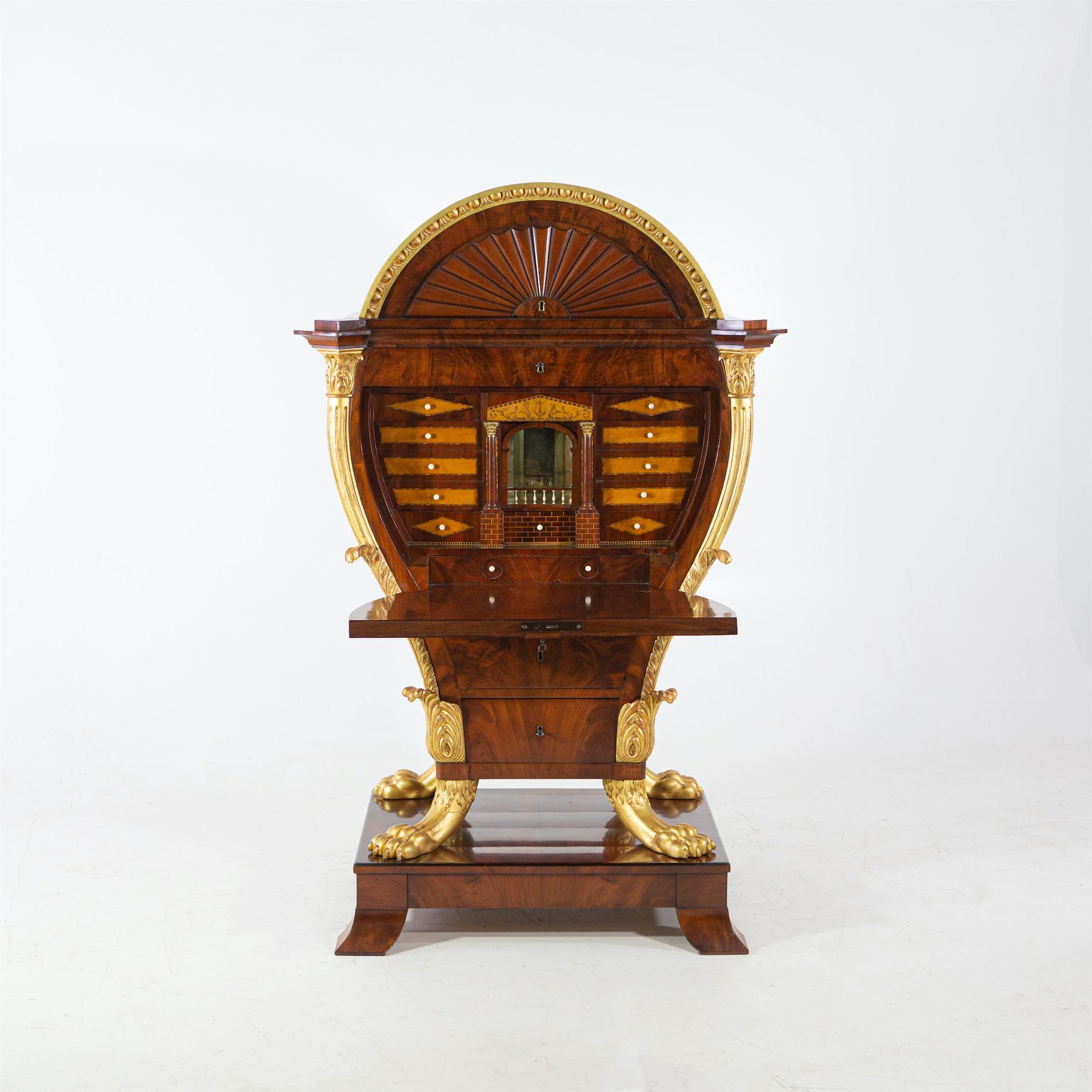 The writing furniture made of mahogany is designed in the shape of a lyre. The gold-plated lion paws stand on a separately manufactured drawer pedestal with square feet curved outwards. The hinged writing compartment with its elaborate interior