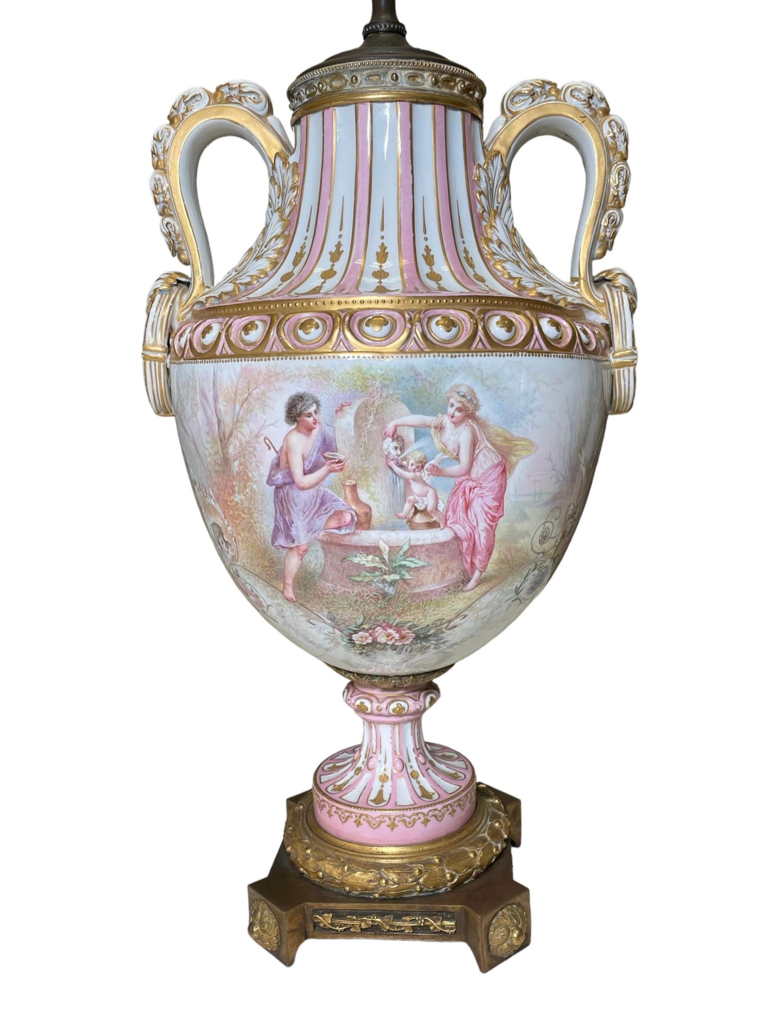 This is a Sevres Style porcelain large bronze mounted urn table lamp. It depicts an urn hand painted pink and white in the background with winged cherubs around the center. Its ear shaped handles are decorated with gilt white scrolls of acanthus