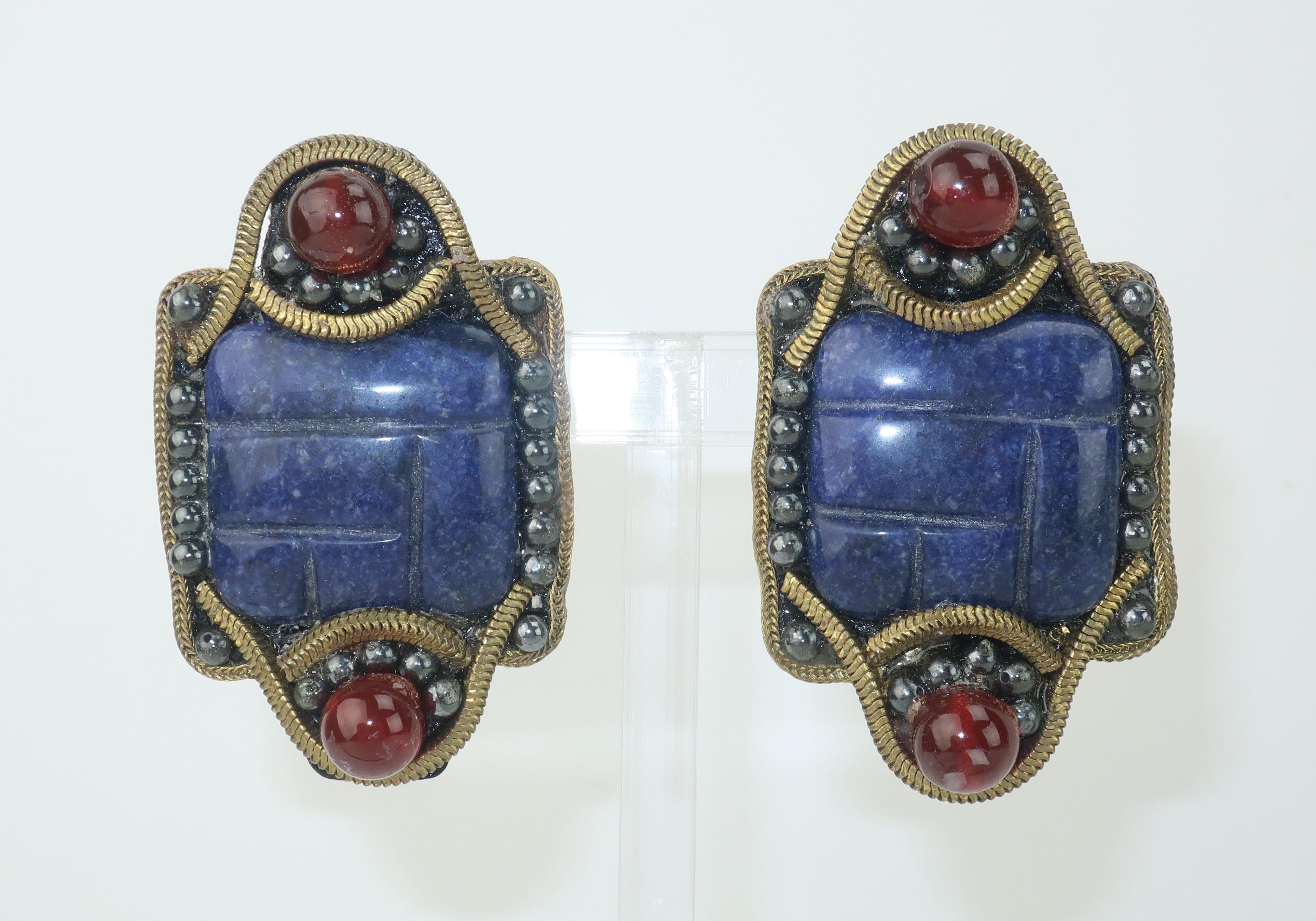 These exotically styled earrings from M & J Hansen are a black resin base accented by blue carved center stones surrounded by carnelian color glass beads and textured chain borders.  Reminiscent of an Egyptian Revival style with a modernist twist. 