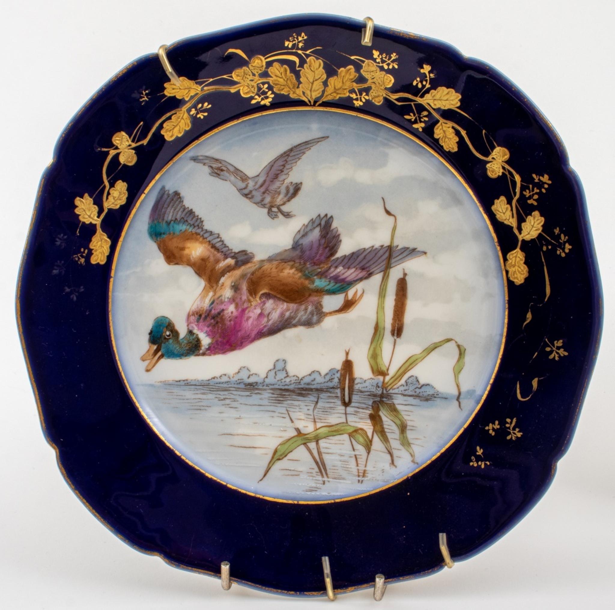 Seven pieces of Limoges ceramic porcelain comprising six plates hand-painted with hunting game including birds, ducks, boars, and rabbits and one service tray with a central vignette of two dogs chasing a stag deer, all with gilt foliate borders on