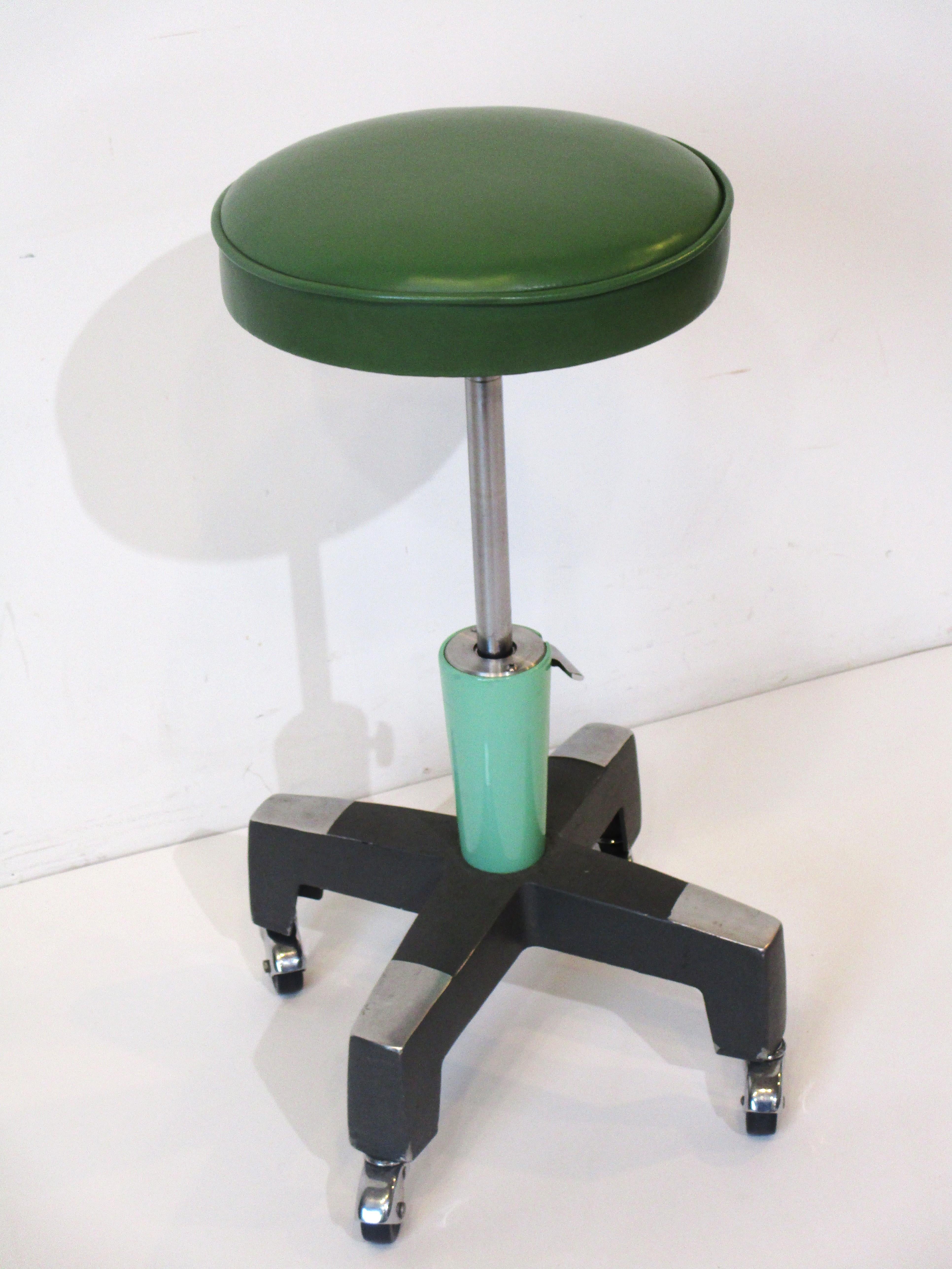 A Machine Age / Art Deco styled rolling adjustable height upholstered stool with green leatherette seat and accents . On a heavy cast steel and aluminum star base with black wheels and chrome wheel fenders having hydraulic adjustments from 19