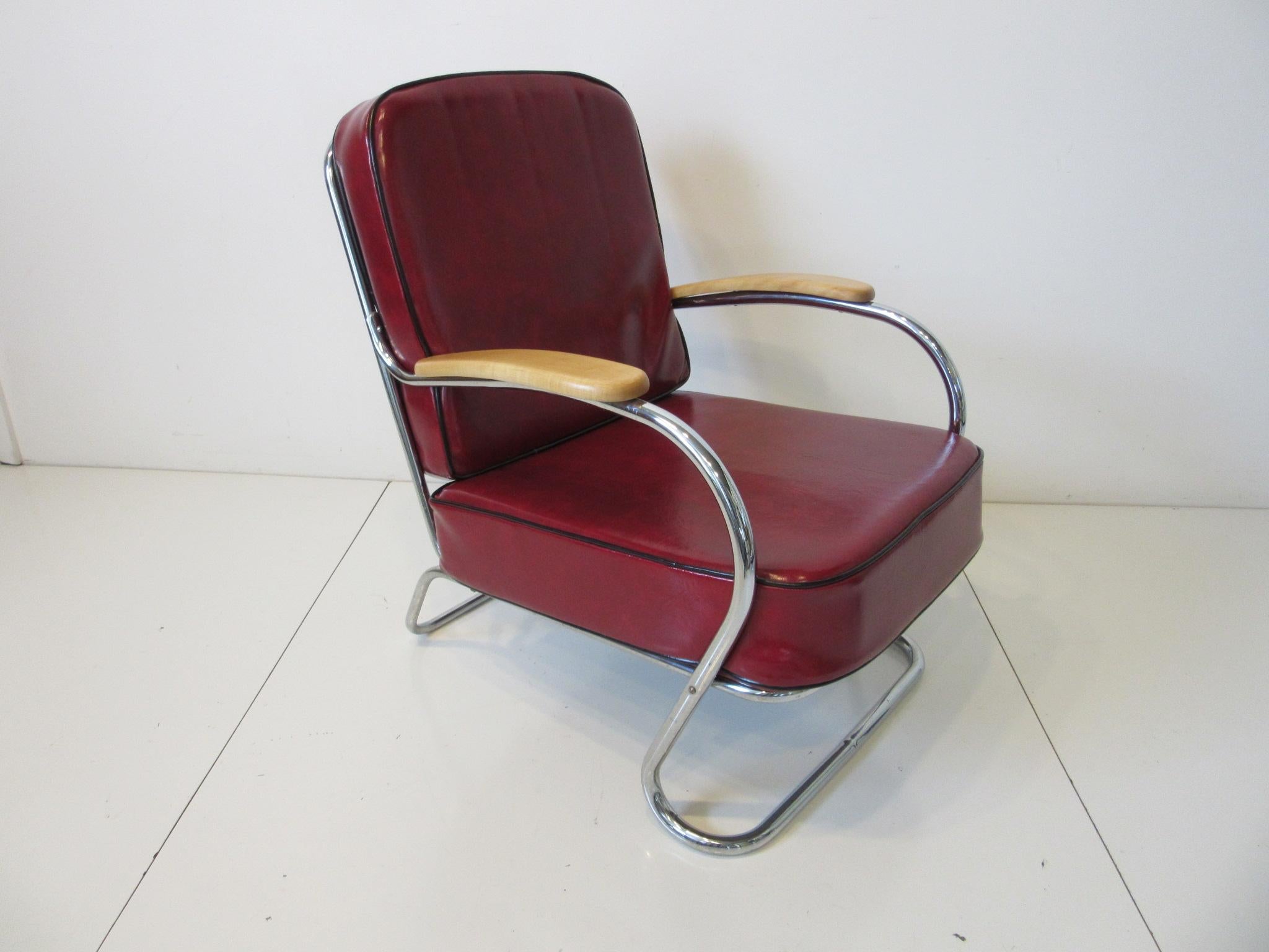 A larger sized Machine Age Art Deco lounge chair with two cushions in red leatherette with black piping sitting on a sculptural chromed steel tubed frame and having wood arm rests. Designed in the manner of Wolfgang Hoffmann and the Howell Furniture