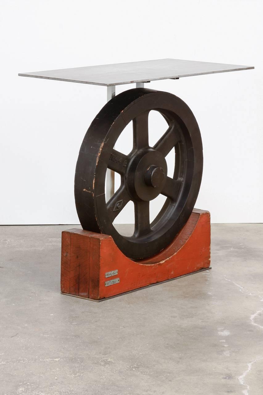 Intriguing Machine Age Industrial art console table featuring a wooden factory wheel displayed on a wood stand surmounted by a steel tabletop. The artistic display is centred by a painted black wheel sitting in a curved red stand. The base has a