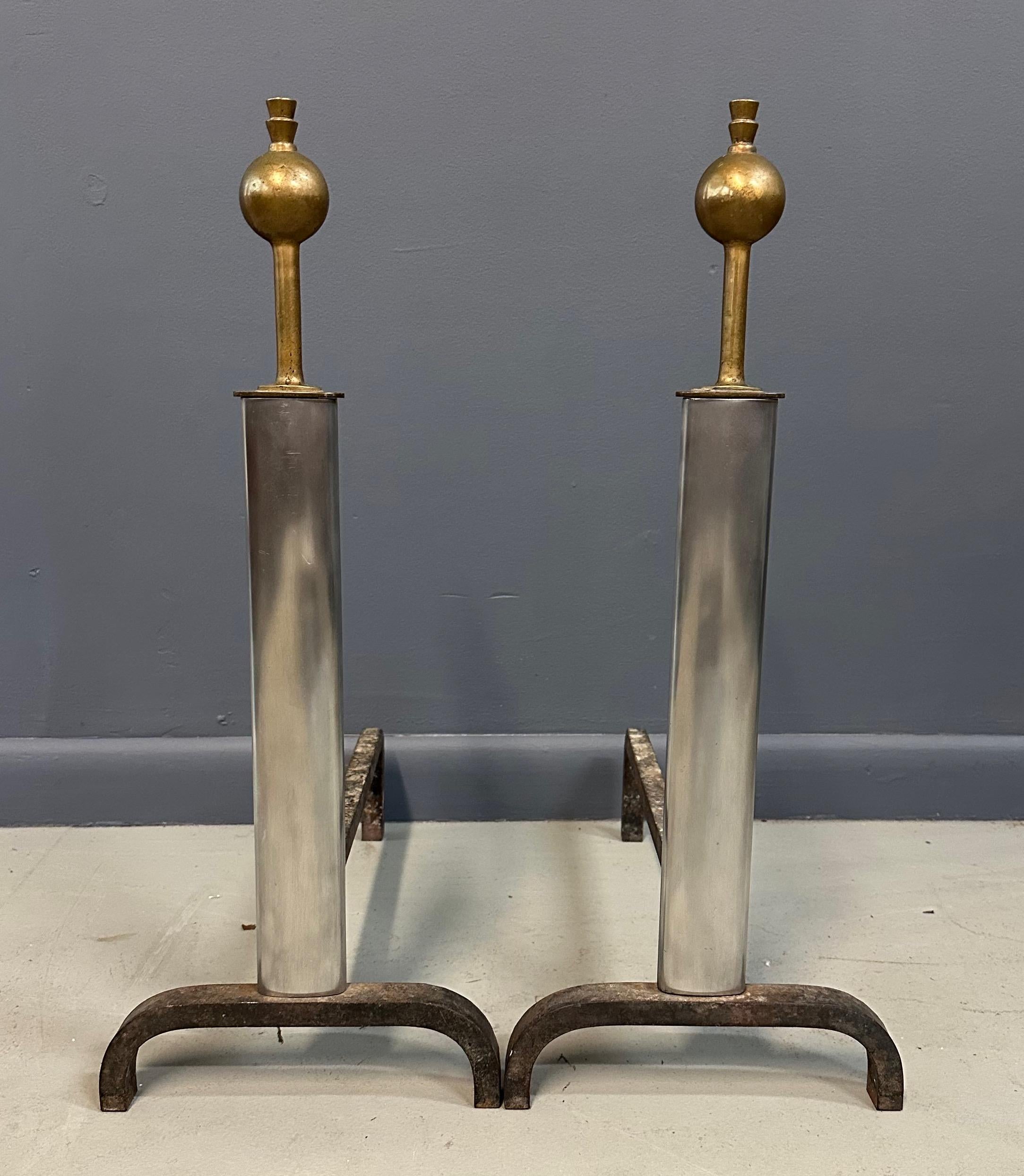 A striking pair of Machine Age andirons featuring a bold industrial aesthetic with contrasting materials. The body is composed of sleek, polished steel columns that rise vertically, capped with sculptural brass finials in a spherical form. The