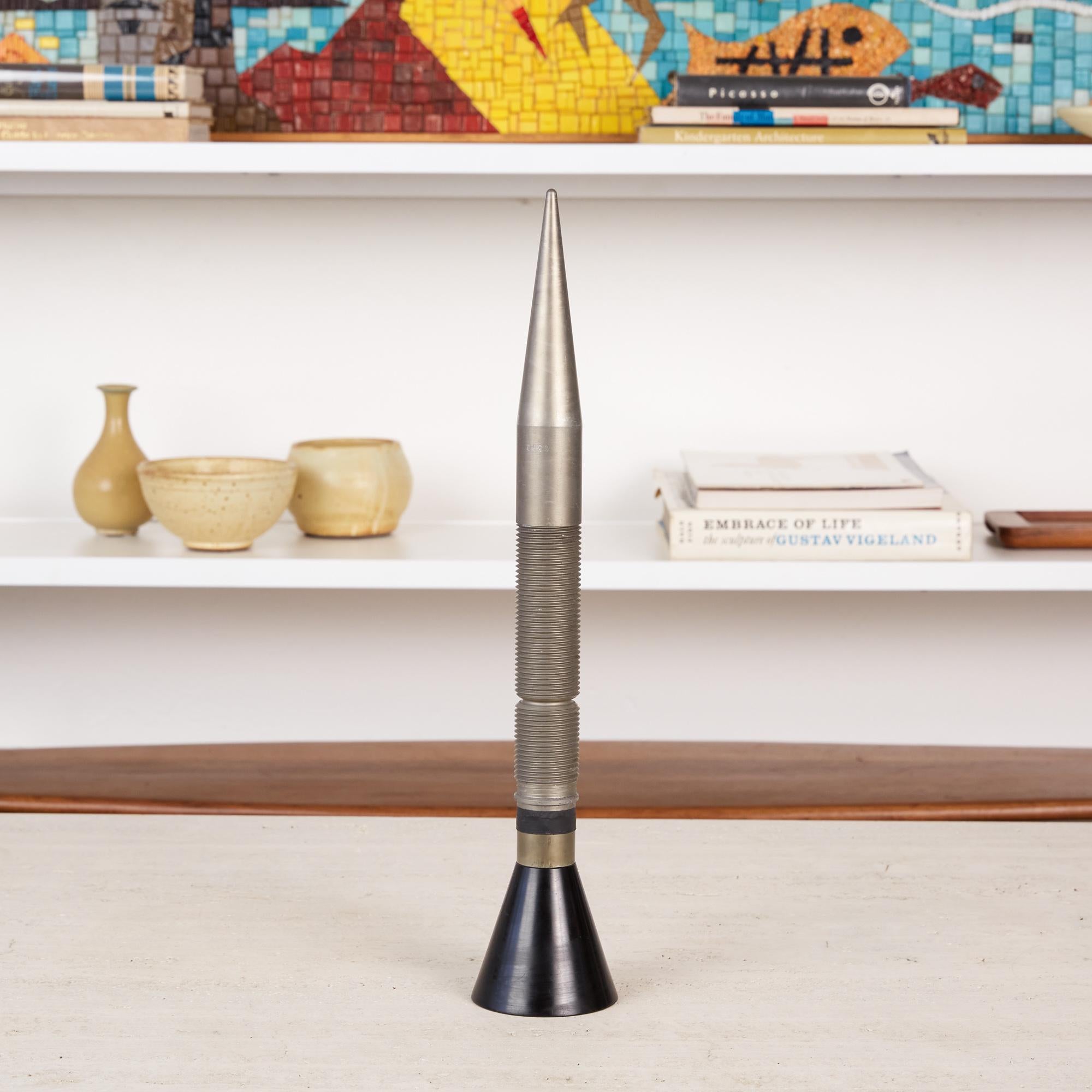 Utilizing components one would typically find in a machinists shop, this abstracted steel rocket is composed of a large steel machine screw that sits atop a black fluted base and has rubber detailing just before the threading begins on the rocket