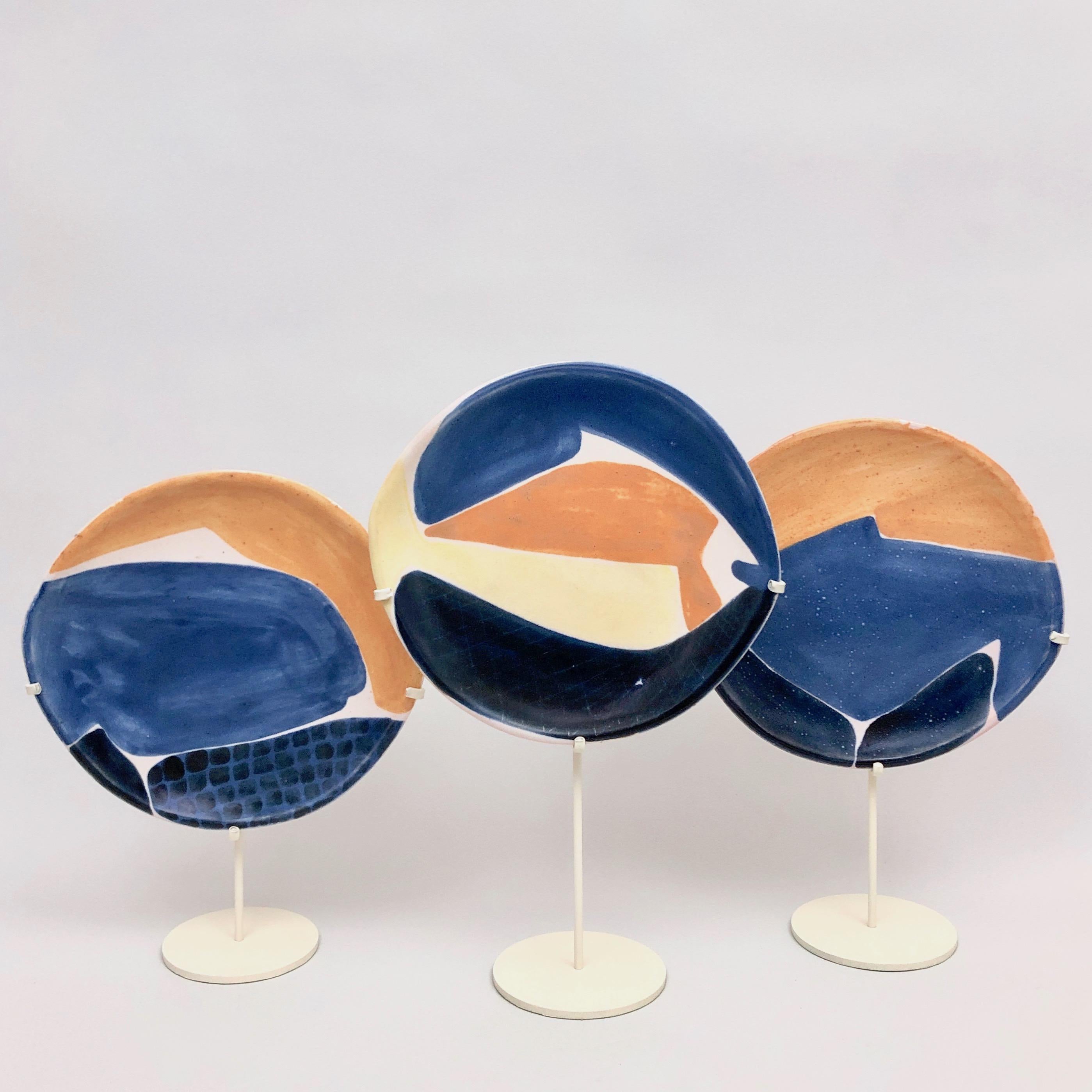 Rare set of 3 ceramic dishes, earthenware glazed in shades of blue, orange, white cream and pale yellow, decorated with designs of stylized fishes. 
The metal bases, with various height dimensions, enameled in white cream, are contemporaries (new),