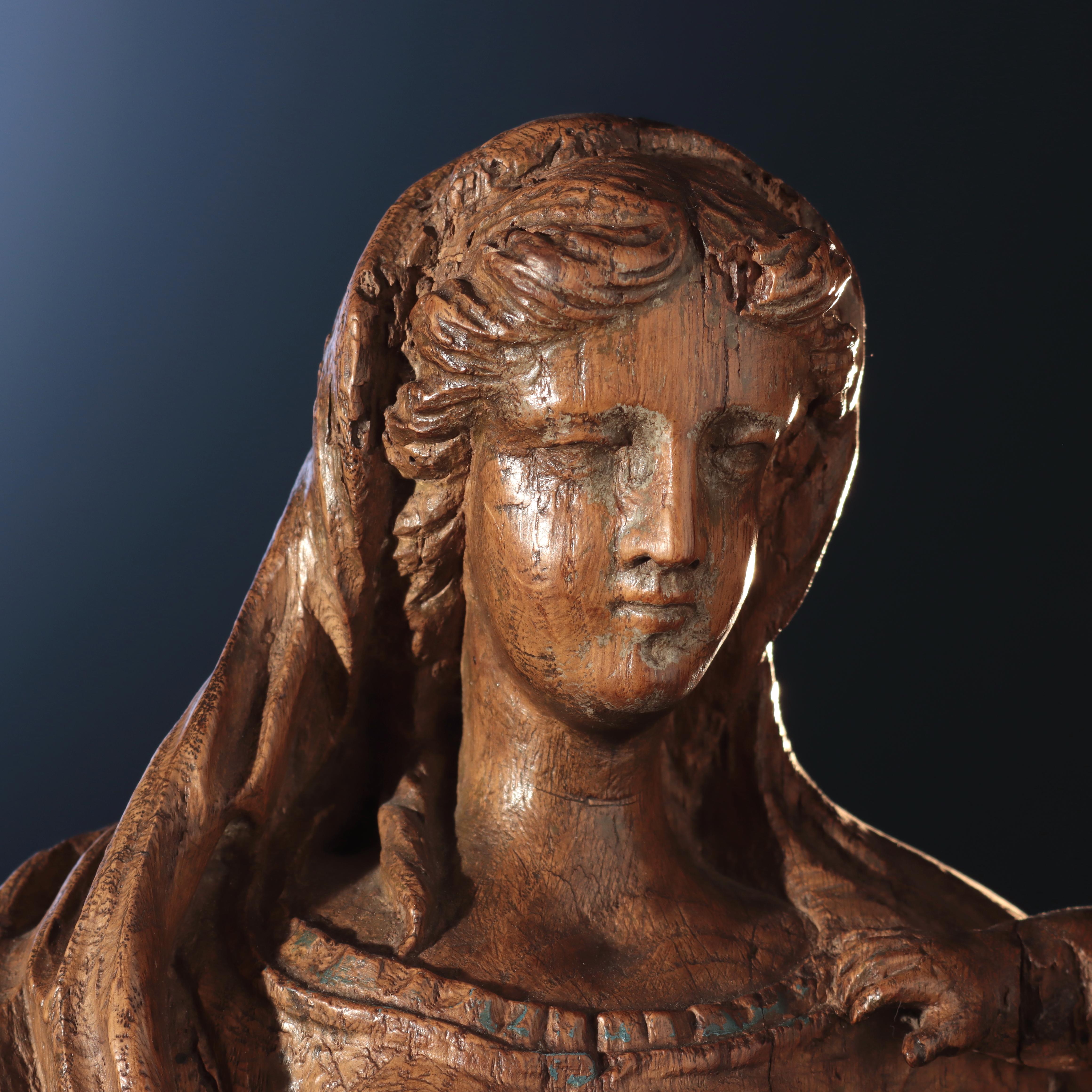 Oak wood sculpture portraying the Virgin standing, her weight on her left leg, to create a spiral movement with her body. She is wearing a vest closed on the front by a tight line of buttons, with a long cape on her head that goes down draping her