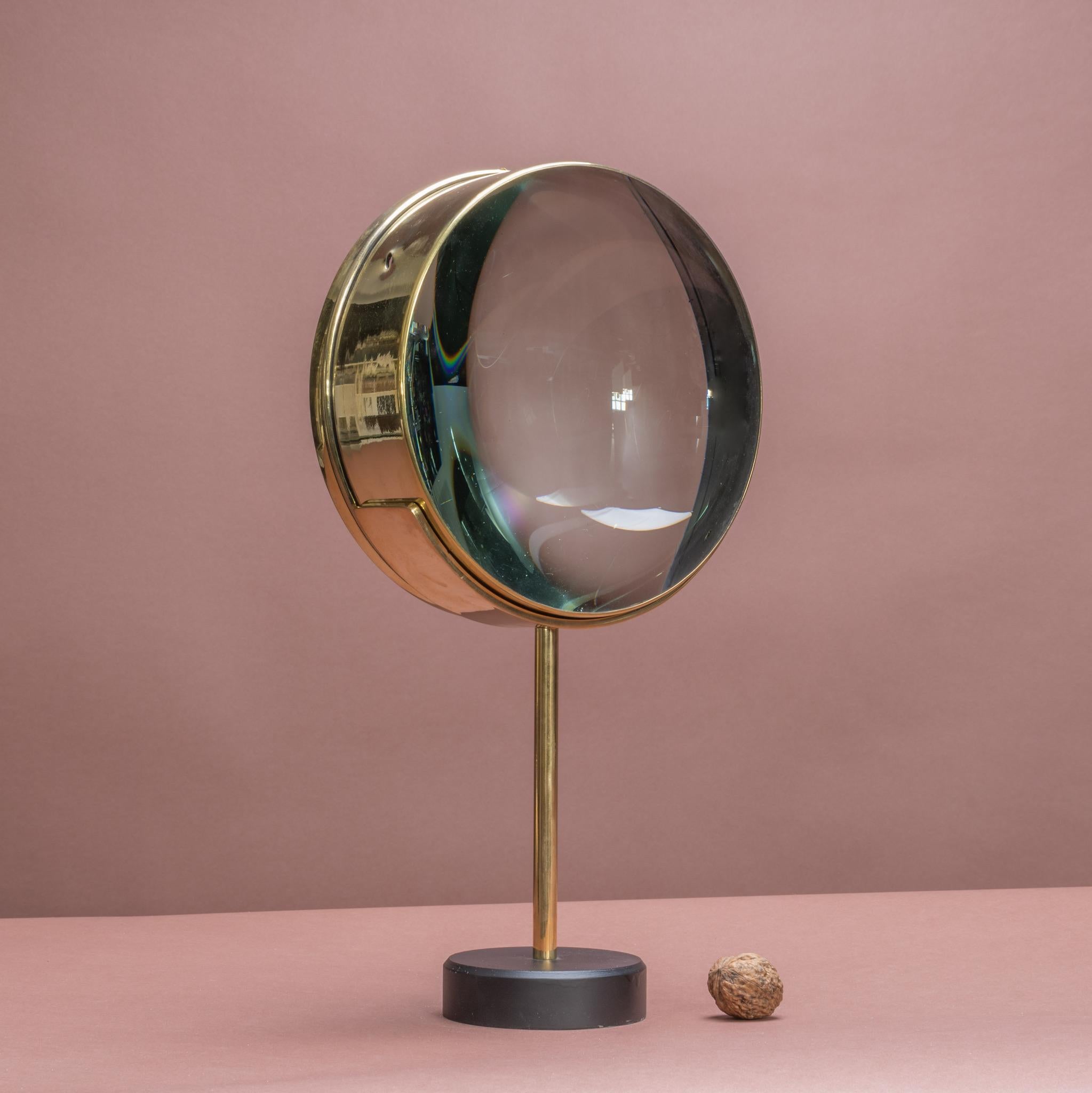 A large lens encased in brass that sits on a newly made display stand. The lens was originally part of a magic lantern which was the earliest form of a slide projector. Magic lanterns were in use from the 18th century until the first quarter of the