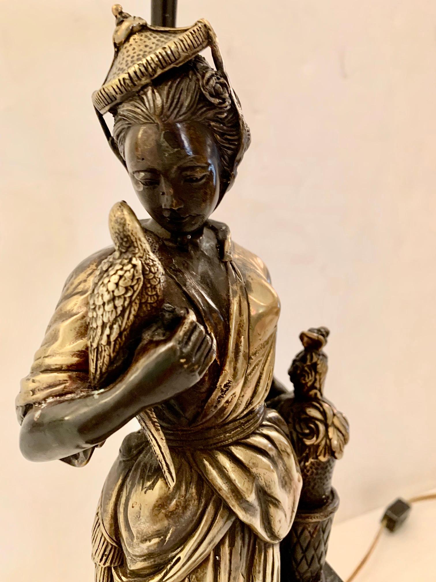 Une belle et inhabituelle lampe de table sculpturale ayant une base figurative asiatique d'une femme aristocratique élégante avec un chapeau à pointe et un oiseau dans une main. La patine est d'un riche argent et bronze tandis que la base est noire.