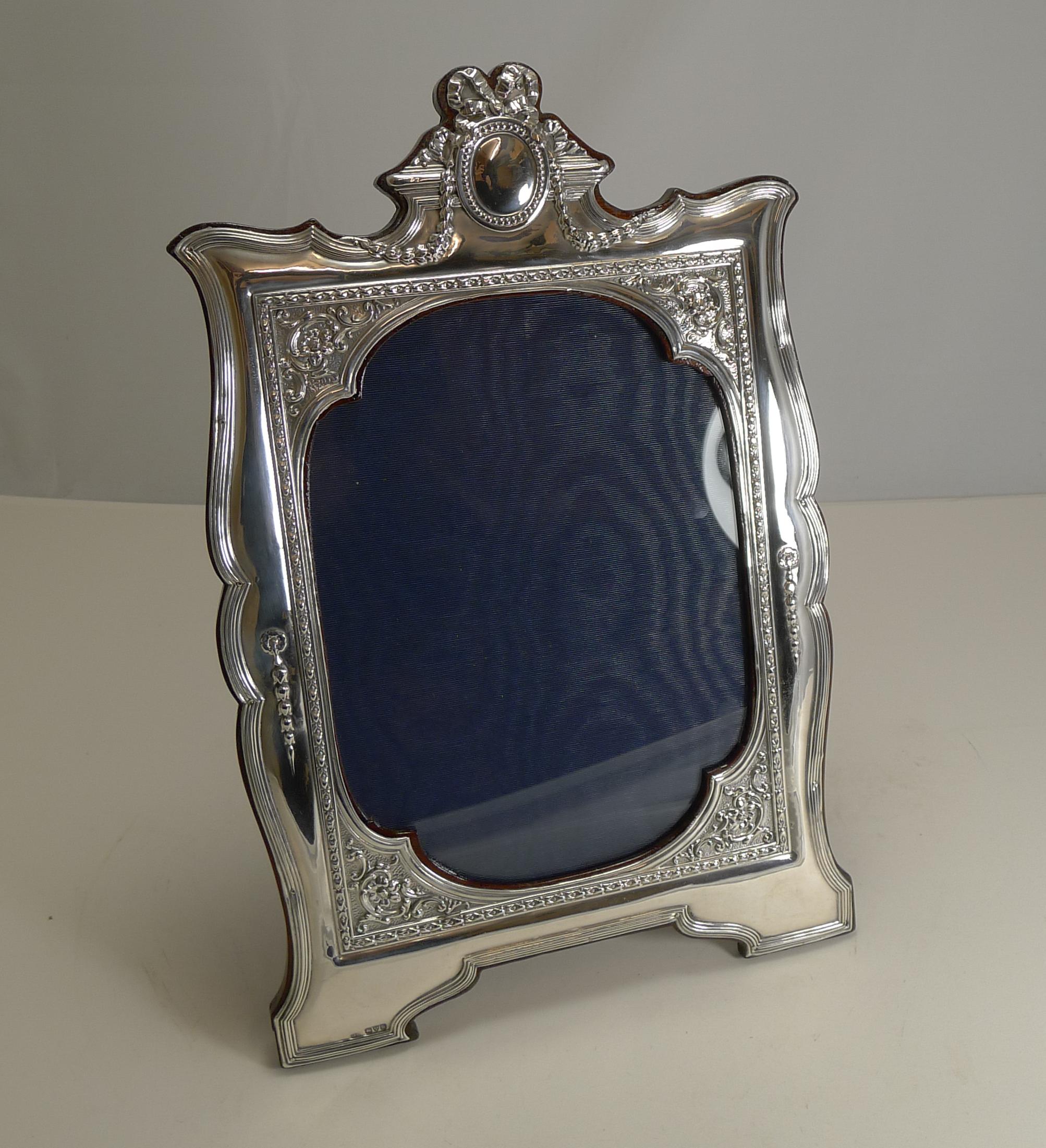 A wonderful large and impressive decorative photograph frame made from English sterling silver with a mahogany backing incorporating a folding easel stand.

The shaped frame is beautifully decorated with a lovely shaped glazed aperture. The silver